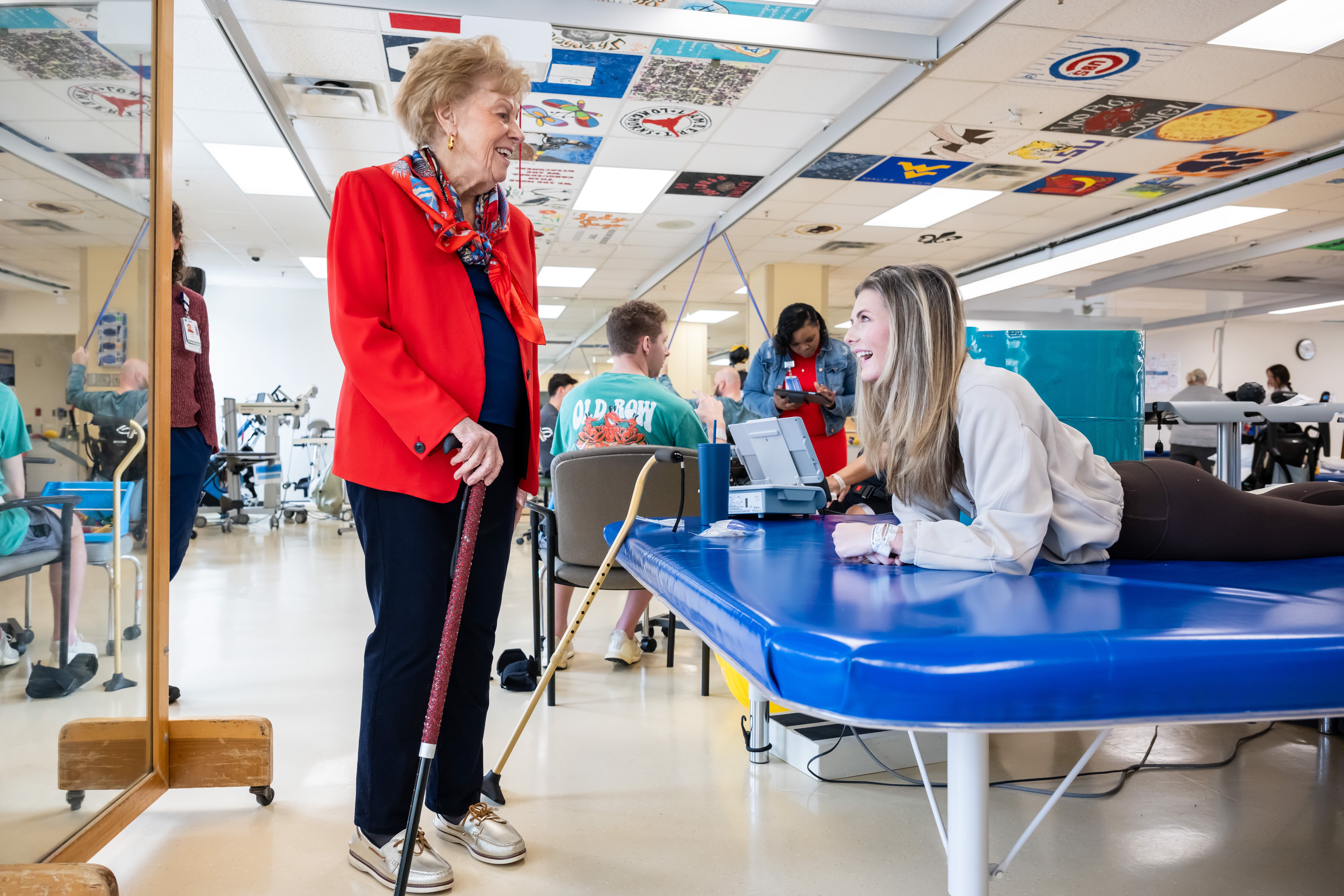 An elderly woman in a red jacket and holding a cane smiles at a young woman lying on a blue therapy table. They are in a rehabilitation facility with colorful ceiling tiles and other people in the background.