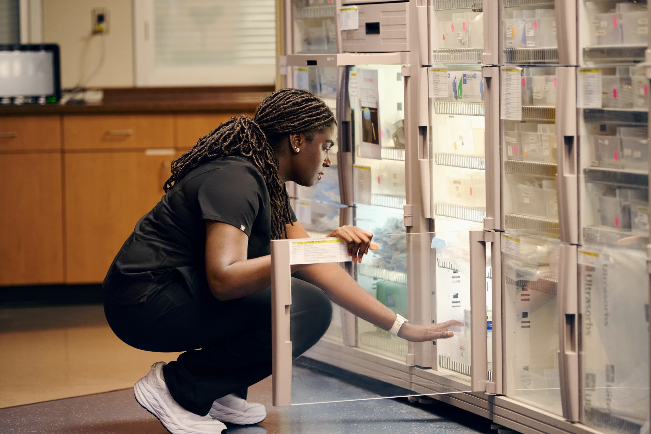 A person wearing black scrubs is crouching and reaching inside a glass-fronted medicine cabinet in a medical or laboratory setting. They have long braided hair and are focused on organizing or selecting items.