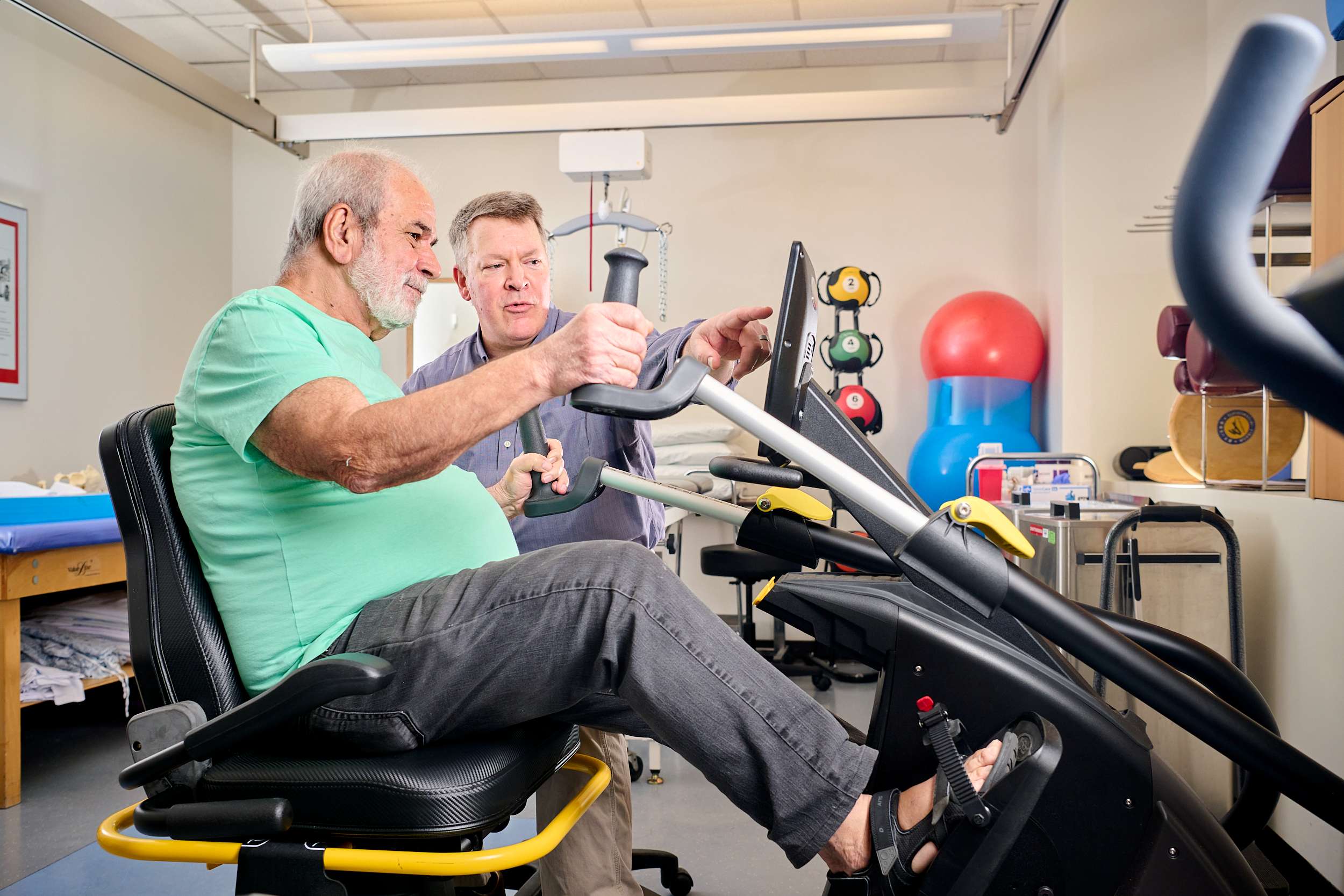 An older man exercises on a stationary bike while a therapist guides him in a rehabilitation room. The room is equipped with colorful exercise balls and other fitness equipment.