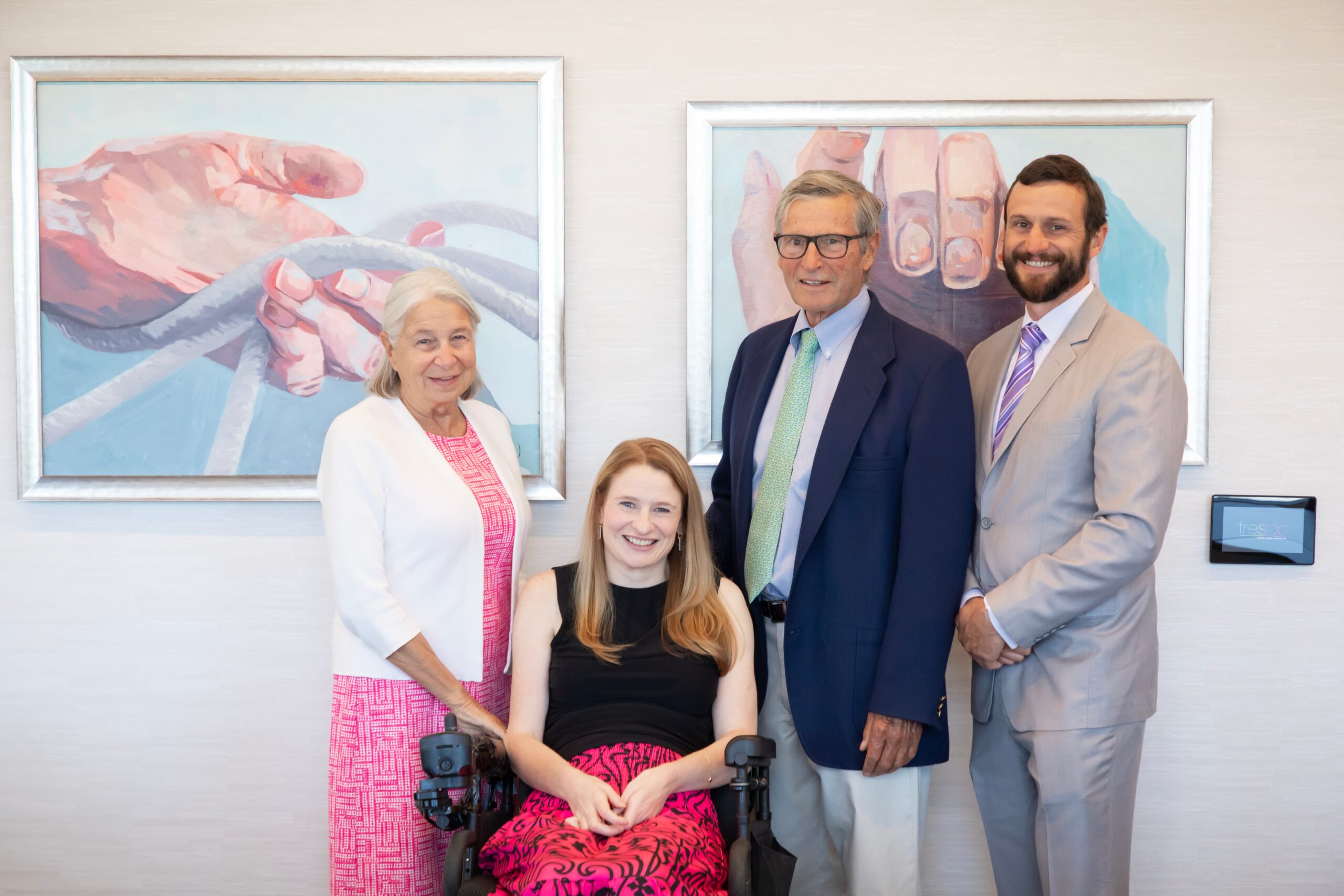 Four people are posing in front of two paintings of hands. A woman in a pink dress, a woman in a wheelchair with auburn hair, a man in glasses with a blue blazer, and a man with a beard wearing a light suit are all smiling.