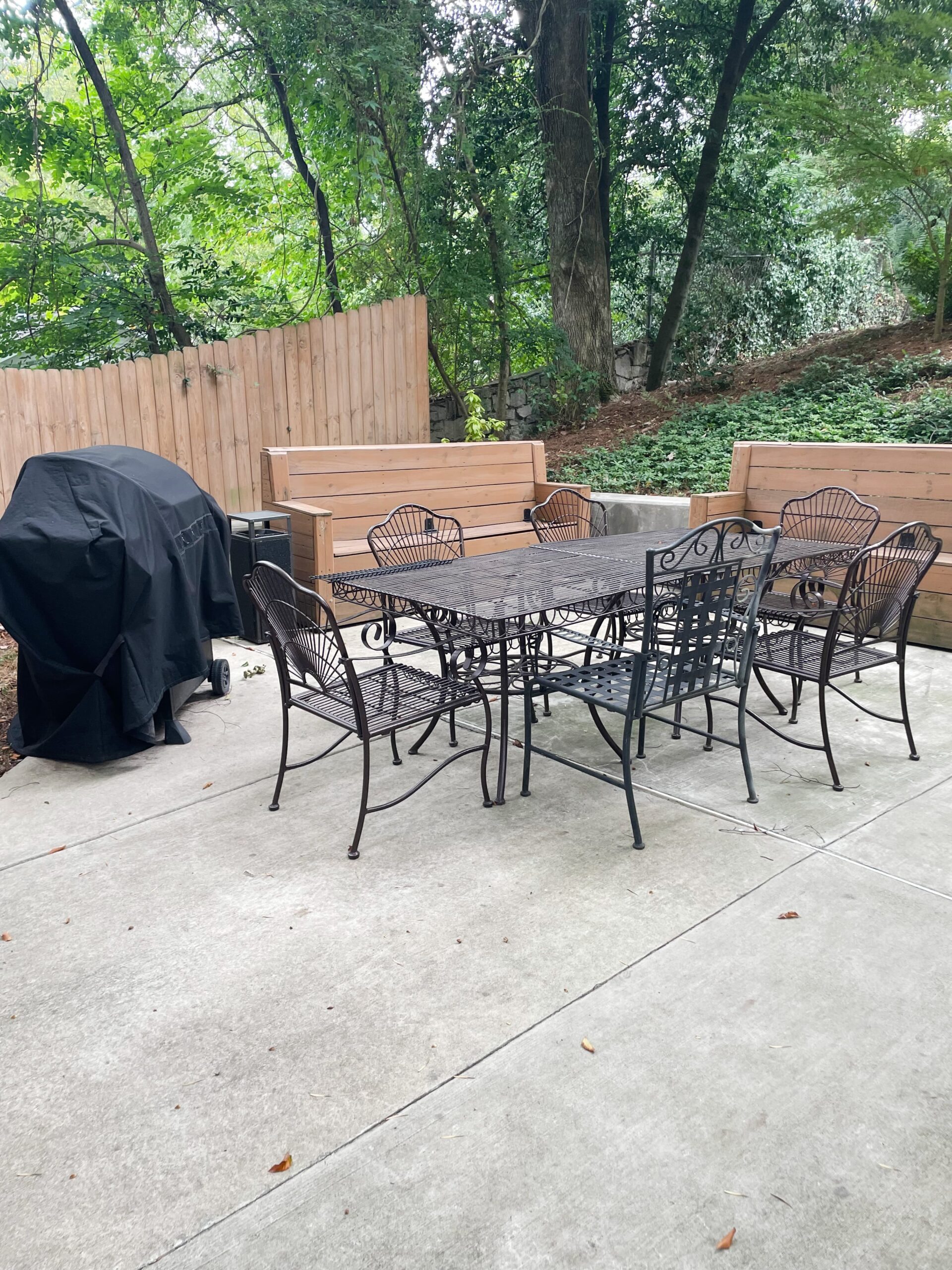 Outdoor patio with a metal dining table and six chairs on a concrete surface. A covered barbecue grill is to the left, and wooden benches are arranged around the perimeter. The area is surrounded by a wooden fence and lush greenery.