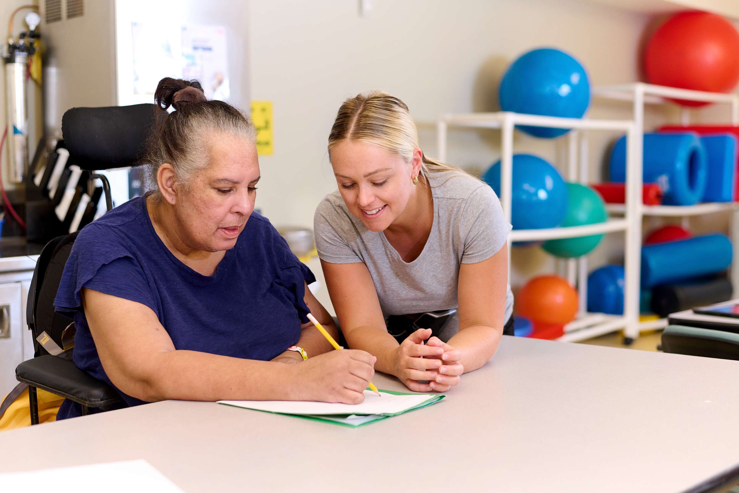A woman in a wheelchair writes in a notebook at a table, assisted by another woman leaning over to help. They are in a room with colorful therapy equipment in the background.