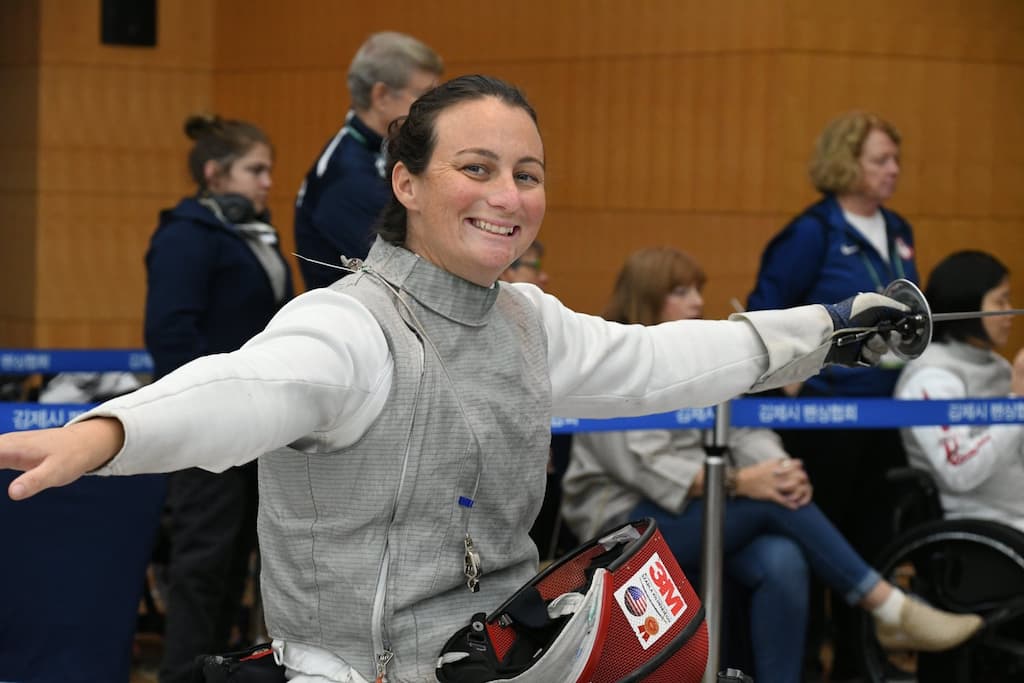 A woman wearing fencing gear and smiling broadly while seated in a wheelchair. She has her arms extended outward. People in the background are watching, and the room has wooden paneling.