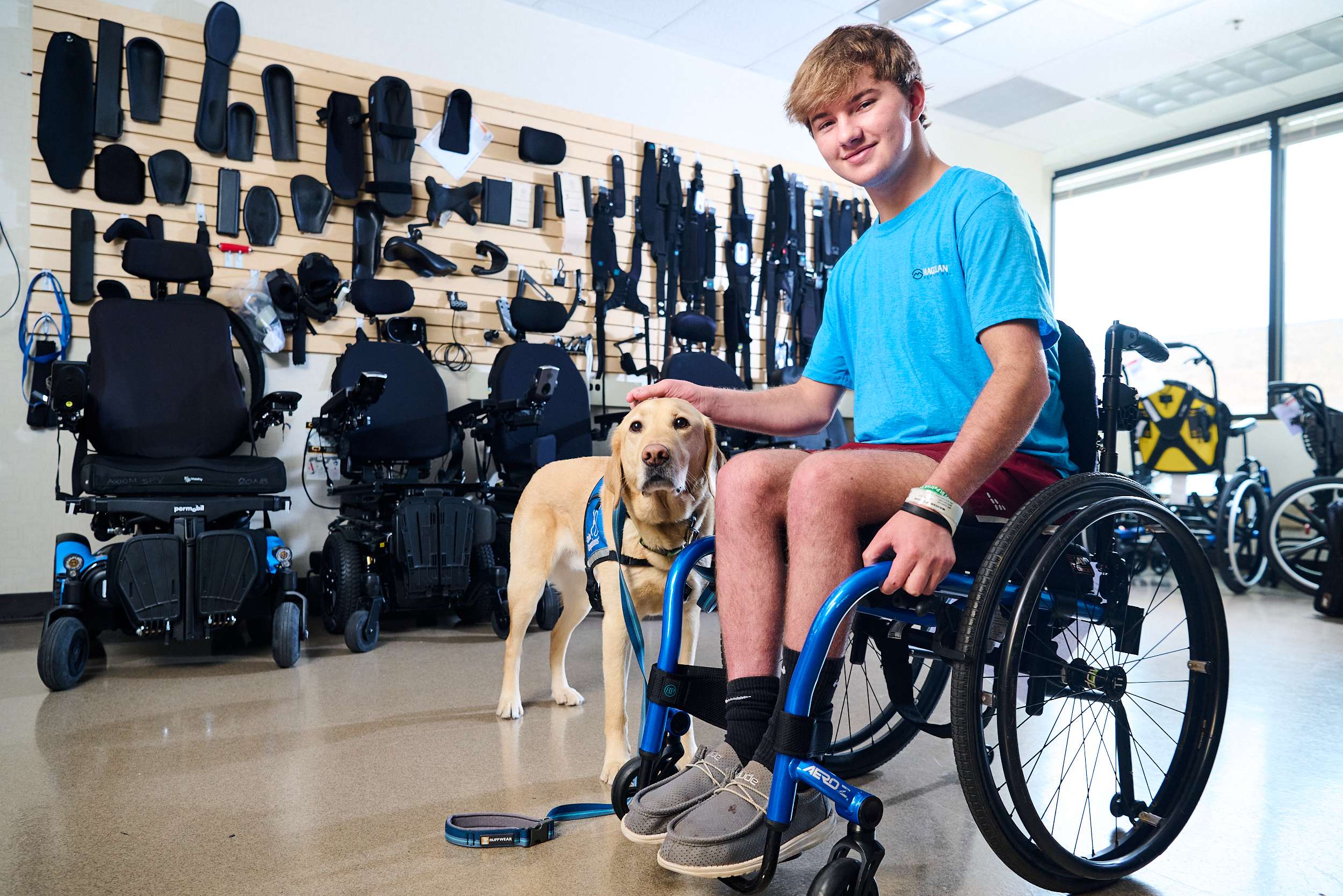 A young person in a blue shirt sits in a wheelchair, smiling and petting a yellow Labrador retriever. Behind them is a display of mobility equipment and accessories on a wall.