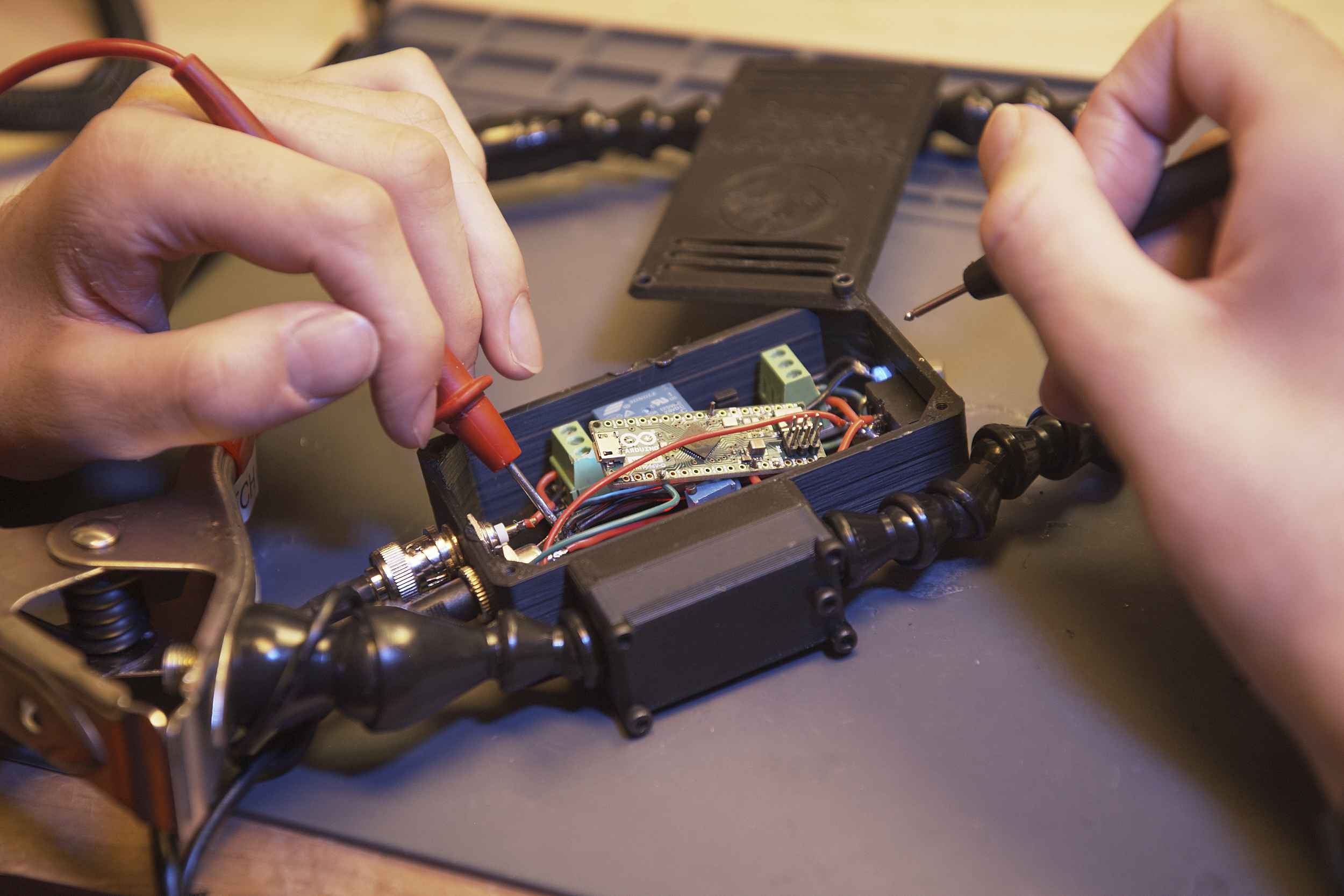 Hands using soldering tools to work on a small open electronic device, with various wires and components visible. The device is held in place by a flexible, adjustable stand on a workbench.