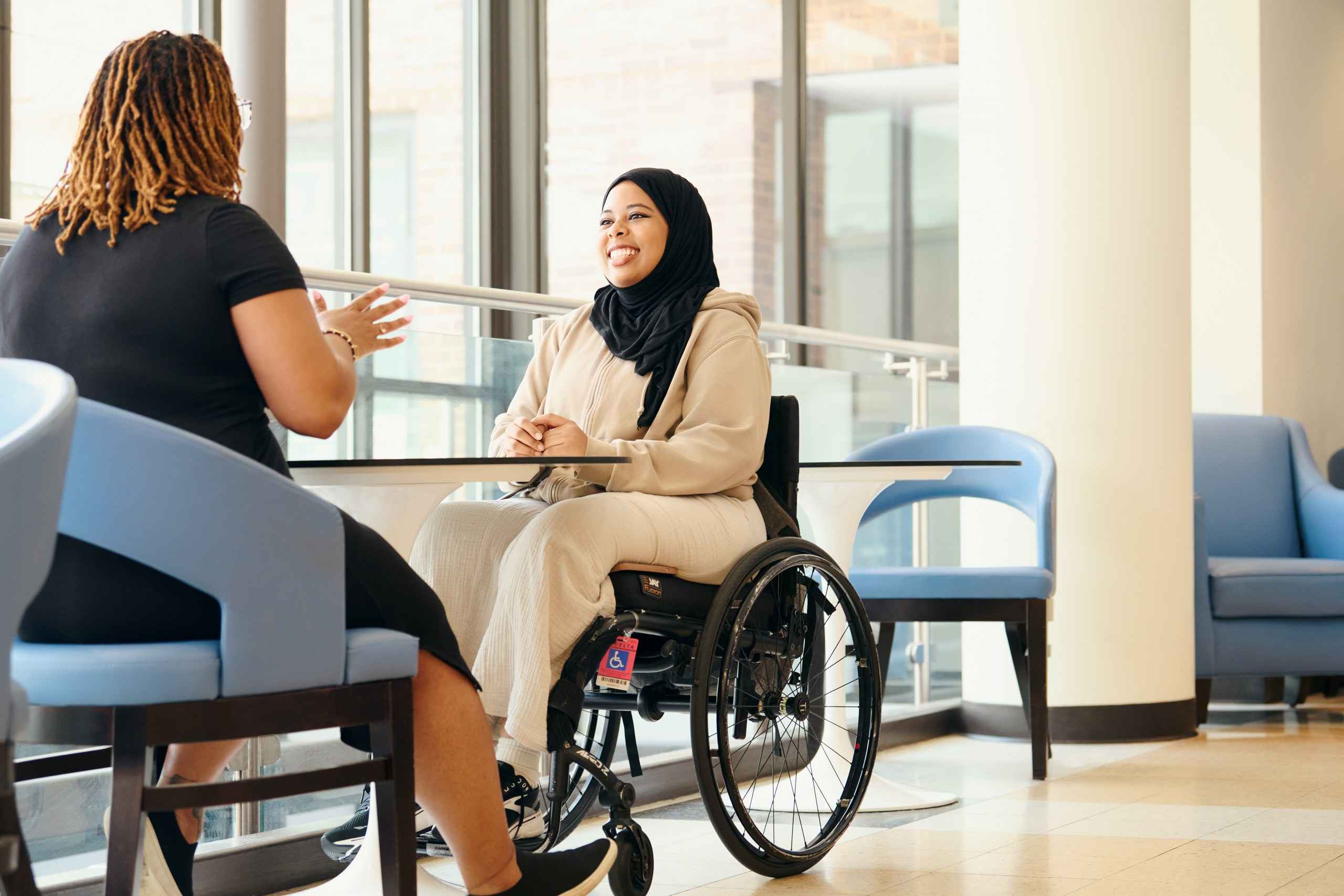 Two women sit across from each other at a table in a bright, modern room. One woman wears a hijab and sits in a wheelchair, while the other gestures as they converse. The space is airy, with large windows and blue chairs.