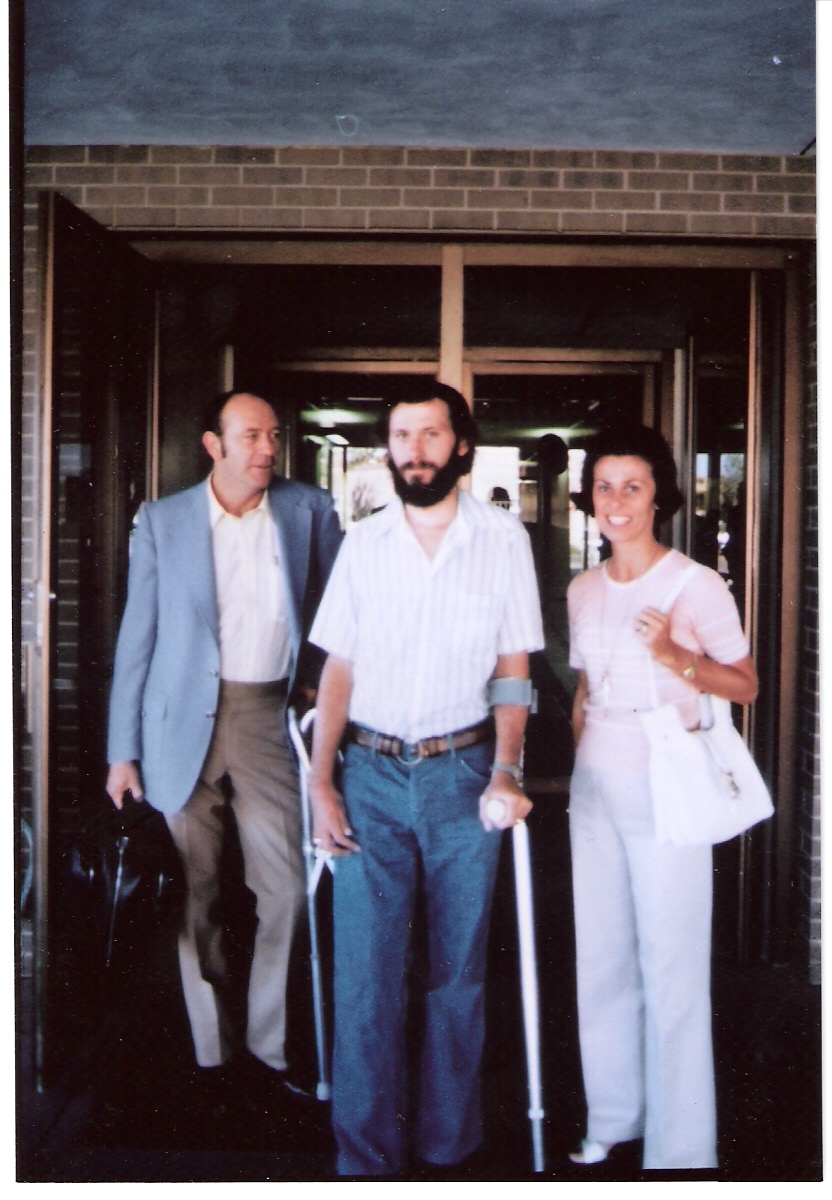 Three people are standing outside a building: Harold Shepherd, James Shepherd, and Alana Shepherd. The person in the middle, with a beard, is using crutches. The person on the left is wearing a light suit, and the person on the right is wearing light casual clothes and holding a white bag.