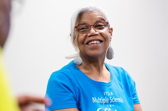 An older woman with short gray hair and glasses smiles warmly. She wears a blue T-shirt that mentions multiple sclerosis and white, large earrings. The background is plain and light-colored.