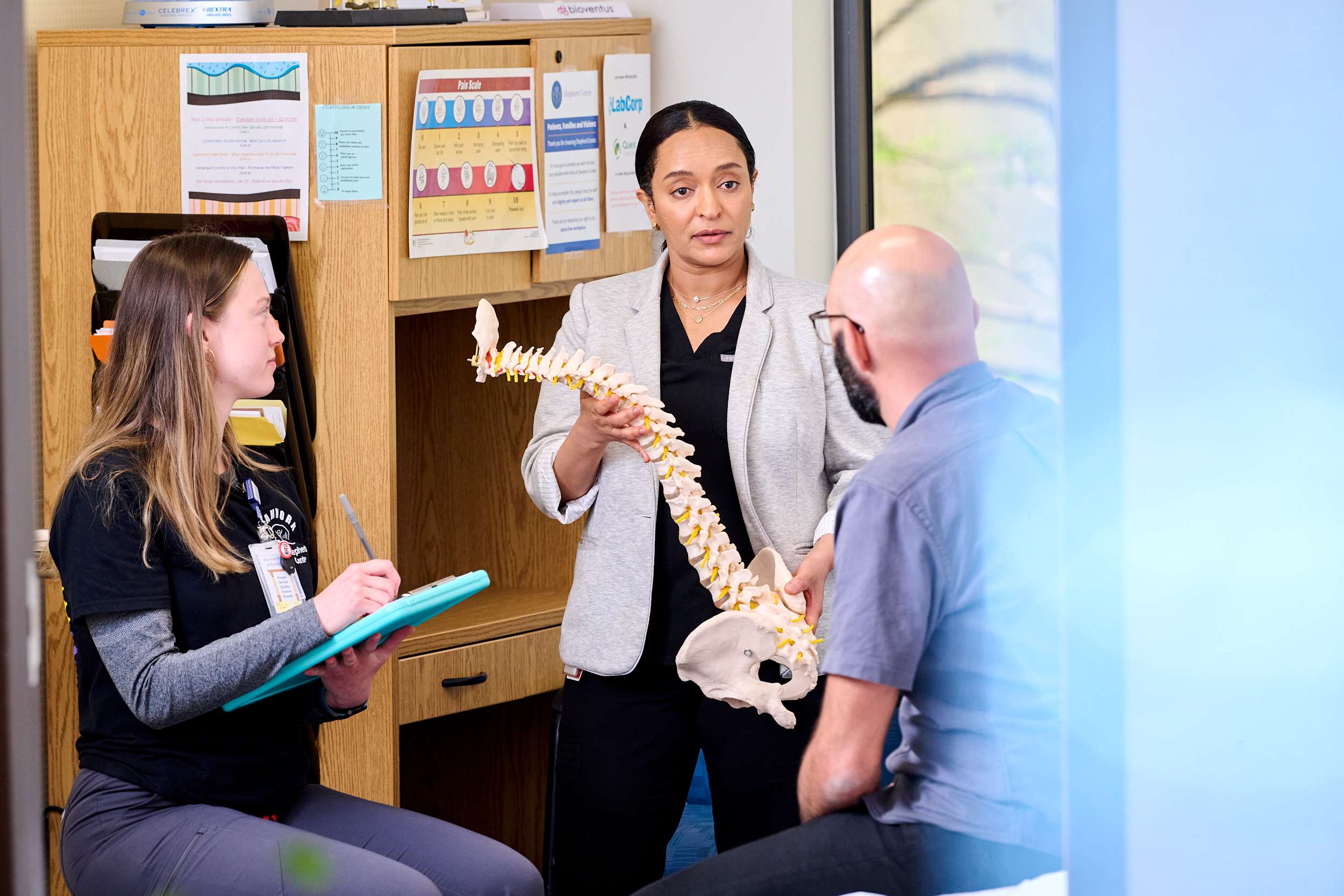 A woman holds a spine model while explaining to a seated man. Another woman sits nearby, taking notes on a clipboard. Shelves with medical charts and posters are in the background.