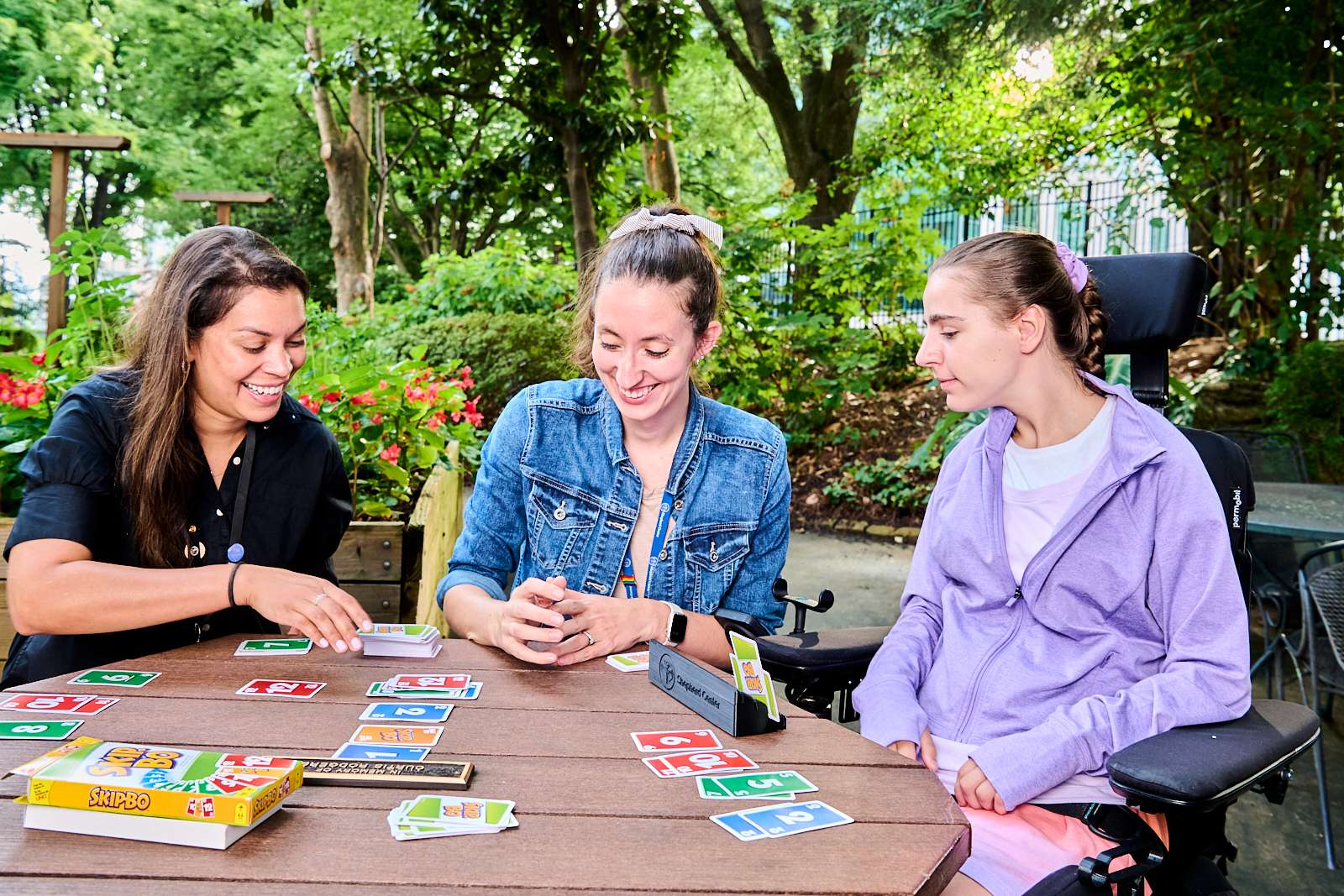 Three people are playing a card game at an outdoor table surrounded by greenery. One person uses a wheelchair. They are smiling and engaged in the game, creating a friendly and inclusive atmosphere.