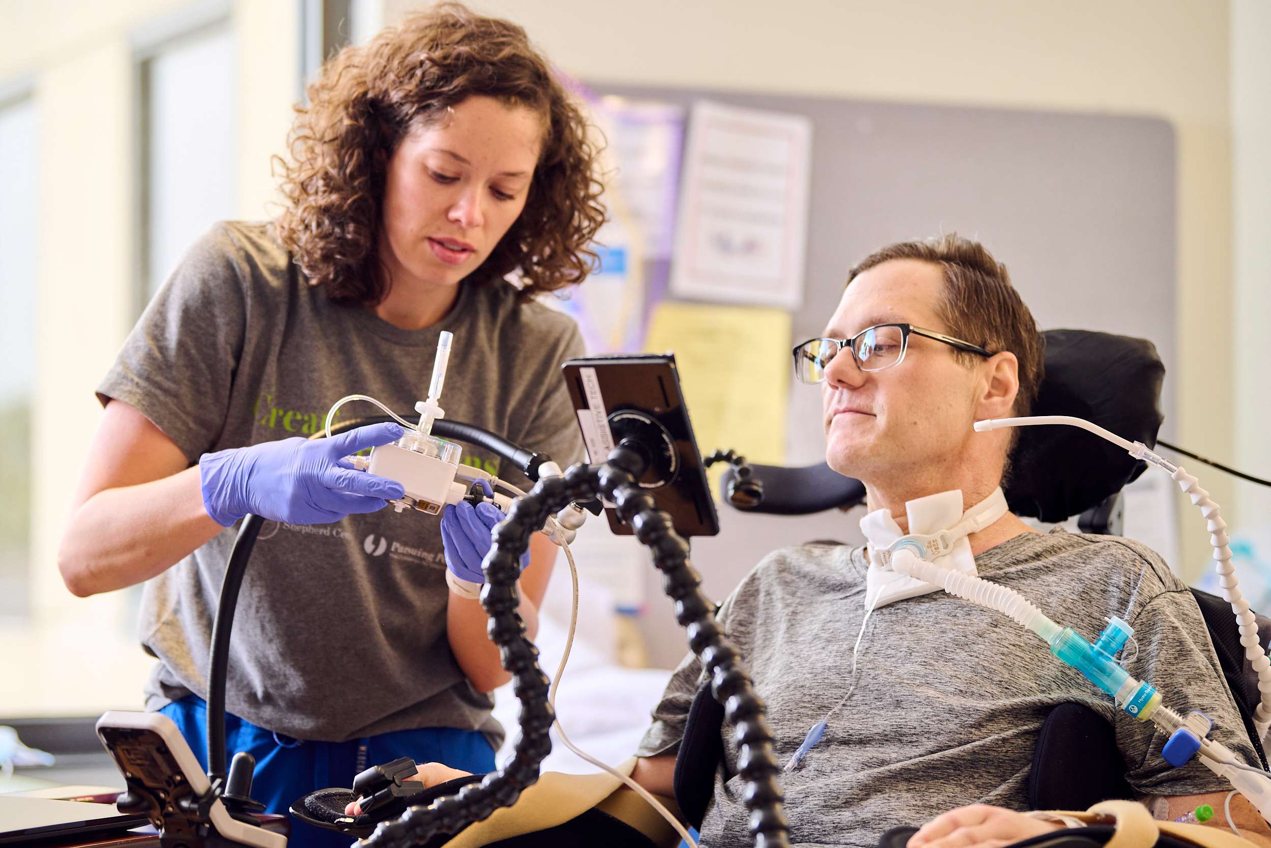 A therapist wearing gloves assists a man using a ventilator in a wheelchair. The man is wearing glasses and looking at a device in front of him. The setting appears to be a bright and supportive care environment.
