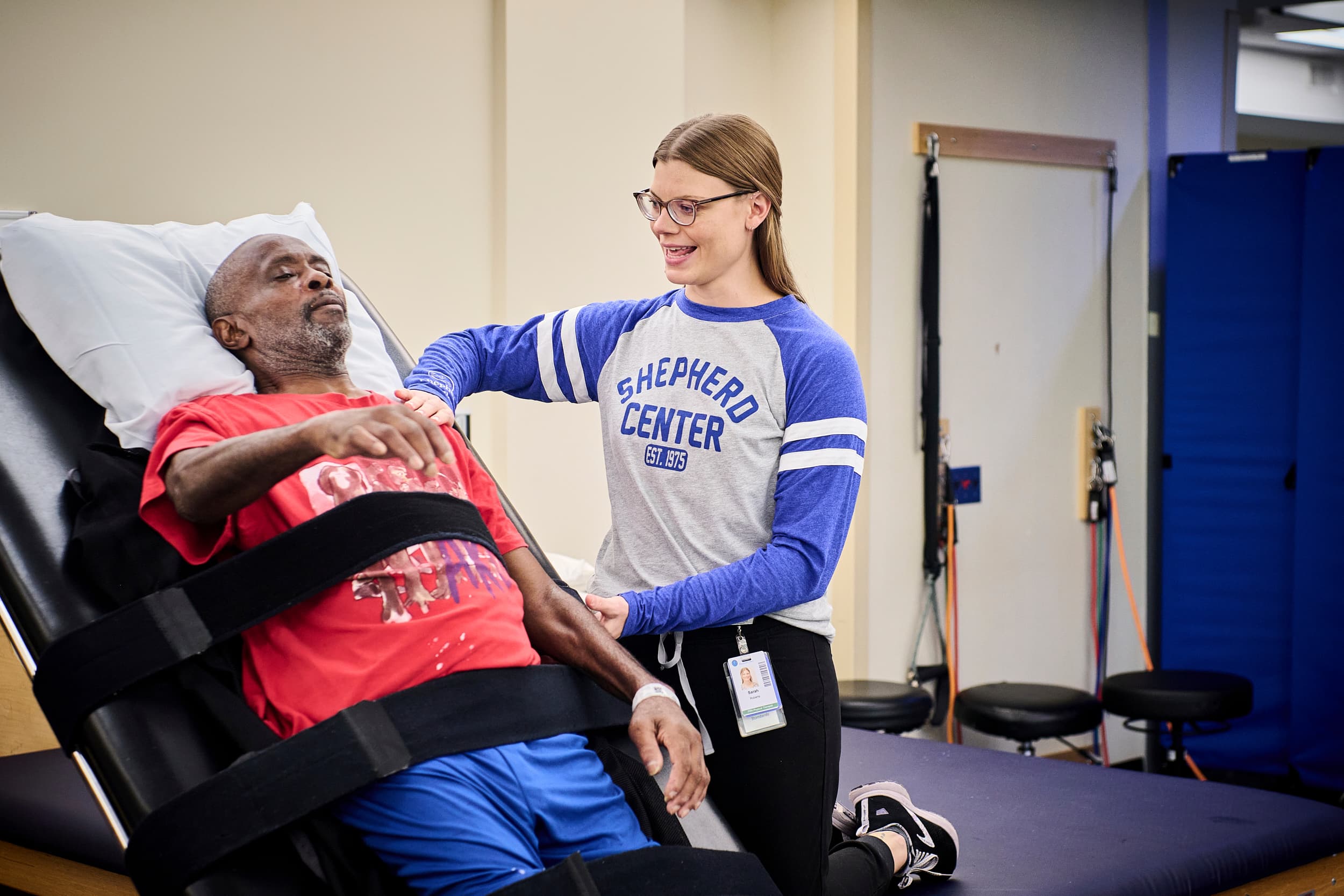 A male patient is reclining on a padded therapy table, secured with straps. A female therapist in a 