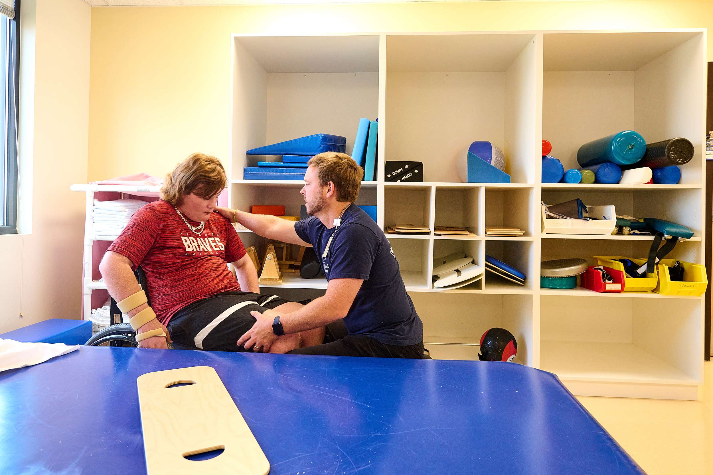 A physical therapist assists a patient seated on a blue mat in a therapy room with shelves filled with exercise equipment. The patient is wearing a red shirt and has wrist supports, while the therapist provides support and guidance.