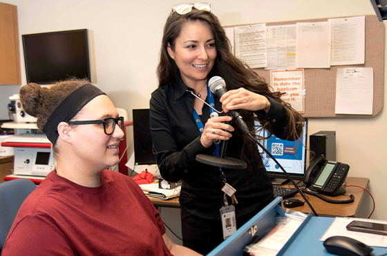 A woman in a red shirt sits at a desk smiling while another woman stands next to her, adjusting a microphone. Papers and bulletin boards are visible in the background.