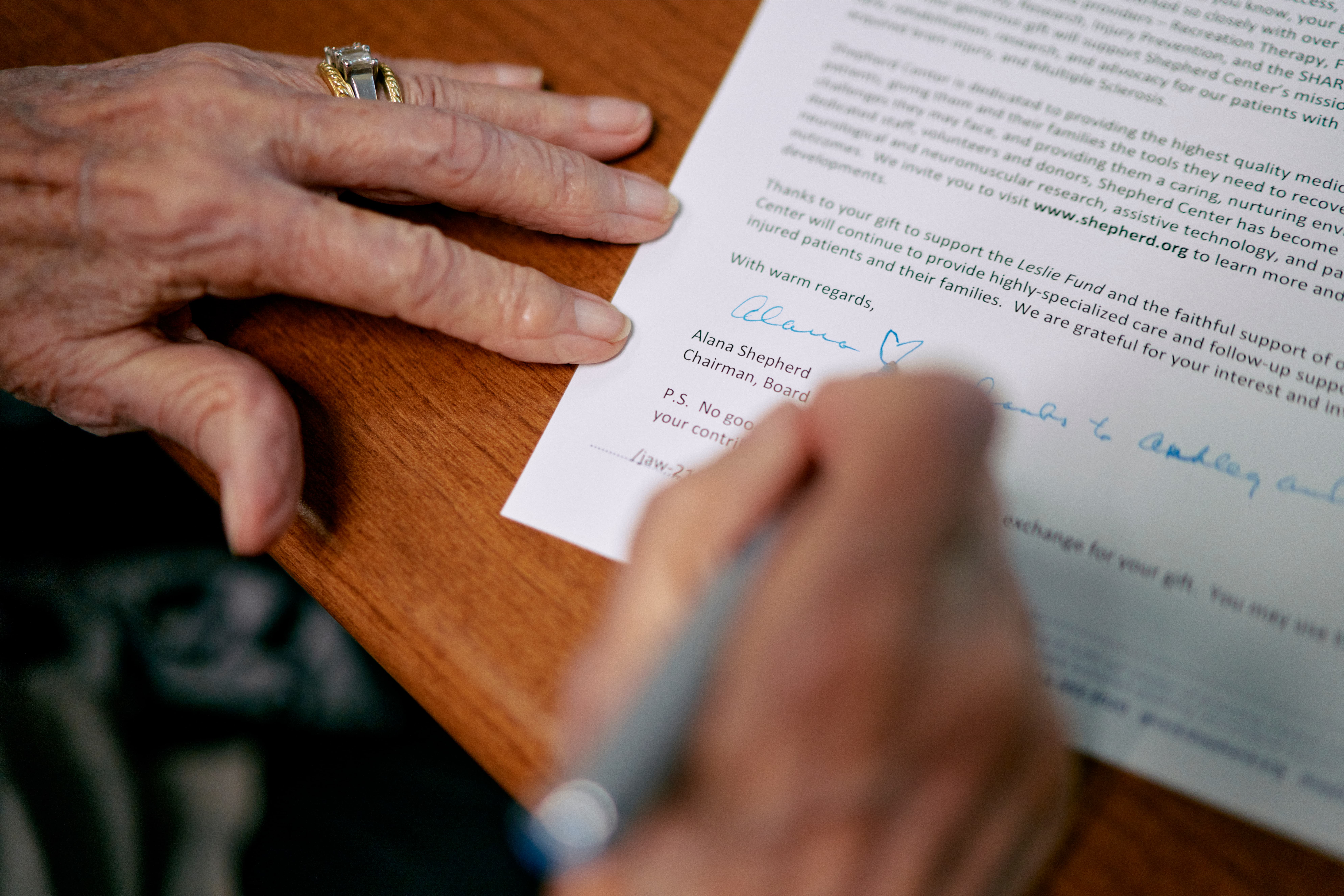 A person with a ring on their finger signs a letter on a wooden table. The letter ends with printed text and a handwritten signature. The page includes a section about supporting a fund and is signed by 