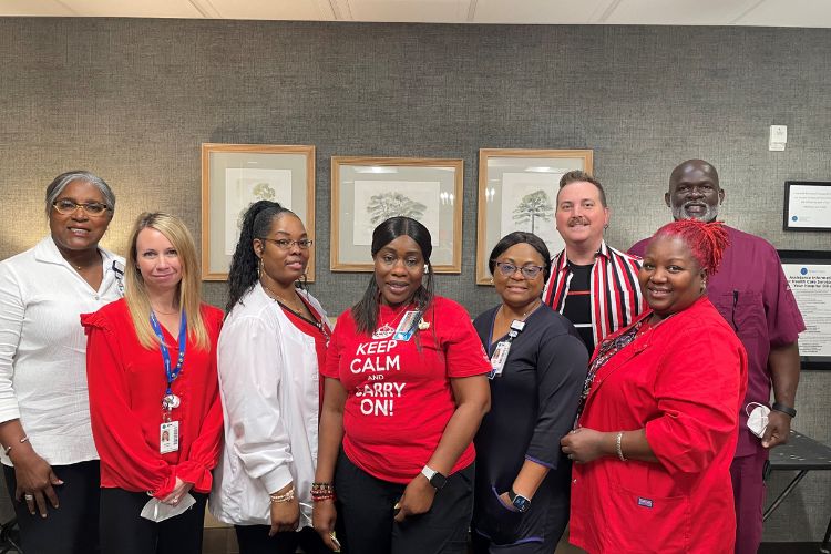 A group of eight people, wearing a mix of red and white clothing, stand together and smile at the camera. They are in an indoor setting with framed pictures on the wall behind them.