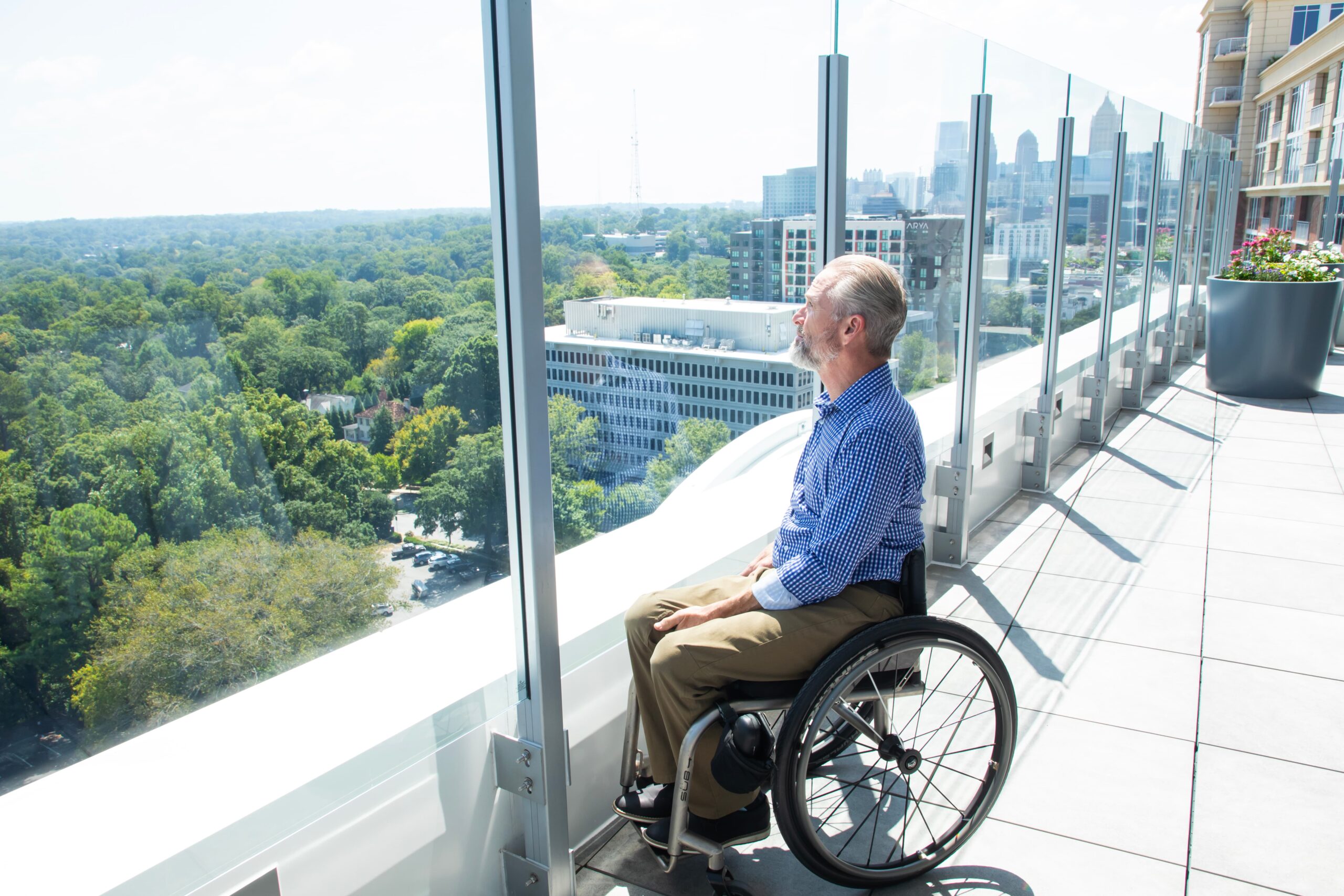 A man in a blue checkered shirt and beige pants sits in a wheelchair on a balcony, gazing at a city skyline and lush green trees. The glass railing offers a clear panoramic view.