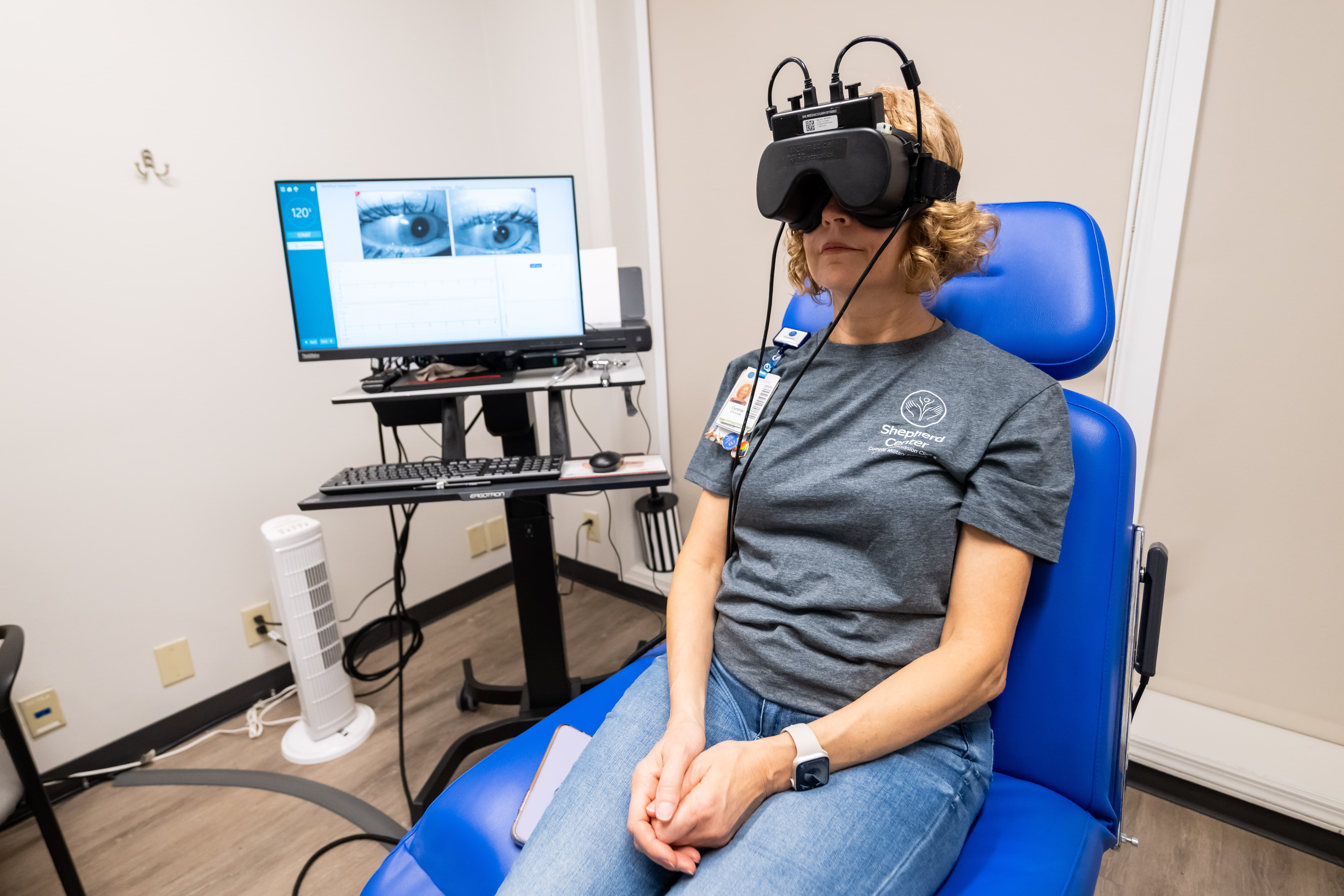 A person sits in a blue chair wearing virtual reality goggles connected to a computer for an eye-tracking study. A monitor displays eye images and tracking data. The person is in a casual outfit with a smartwatch on their wrist.