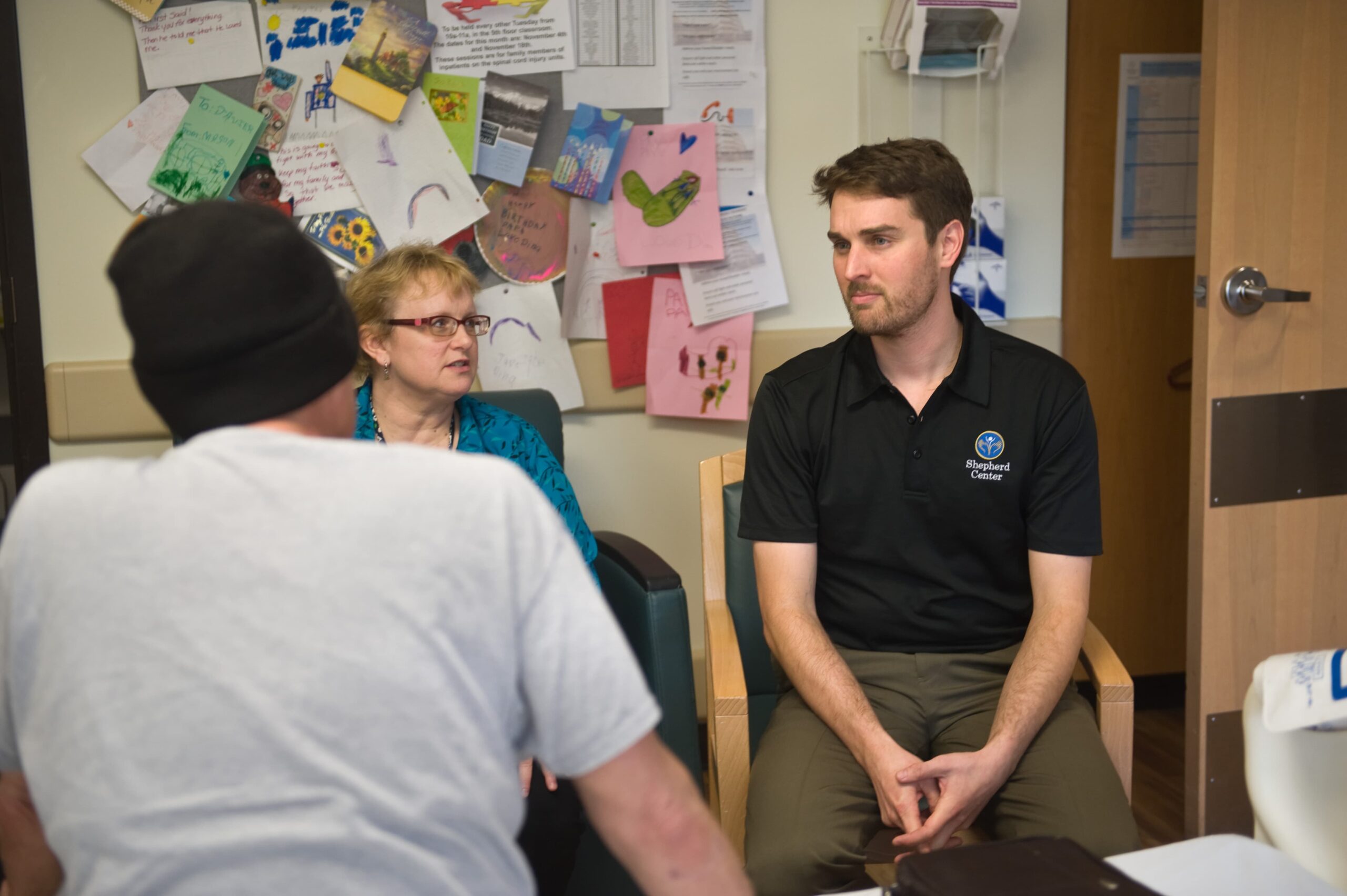 A man in a beanie talks to a woman and a man in a black shirt seated in a room. The walls are covered with colorful drawings and notes. There is a closed wooden door in the background.