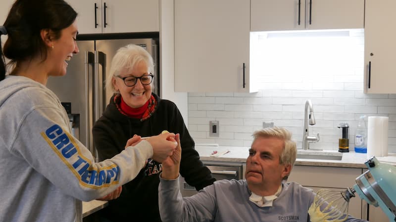 A young woman hands a baking item to an elderly man, who is recovering from Guillain-Barré syndrome, as he sits at a kitchen table. Beside him, an older woman stands smiling. The kitchen boasts white cabinets and a light-colored backsplash.