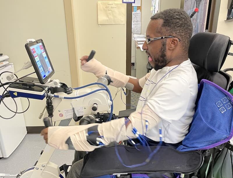 A person, who may be recovering from Guillain-Barre syndrome, sits in a wheelchair using exercise equipment with arm supports and a monitor. They wear protective sleeves on their arms and appear deeply engaged in a physical therapy session.