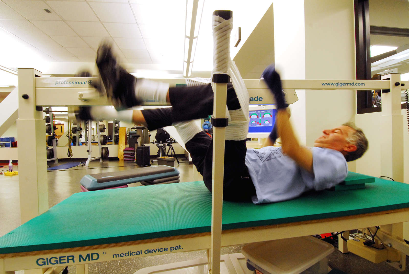 An older adult exercises on a specialized fitness machine in a gym. He is lying on his back with his legs elevated and hands holding onto the equipment. The gym is equipped with various fitness machines in the background.