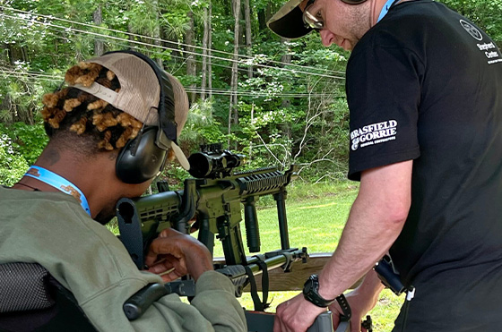 Two people at an outdoor shooting range. One person in a wheelchair aims a rifle with the assistance of the other person standing nearby. Both are wearing ear protection and caps. Trees and greenery are visible in the background.