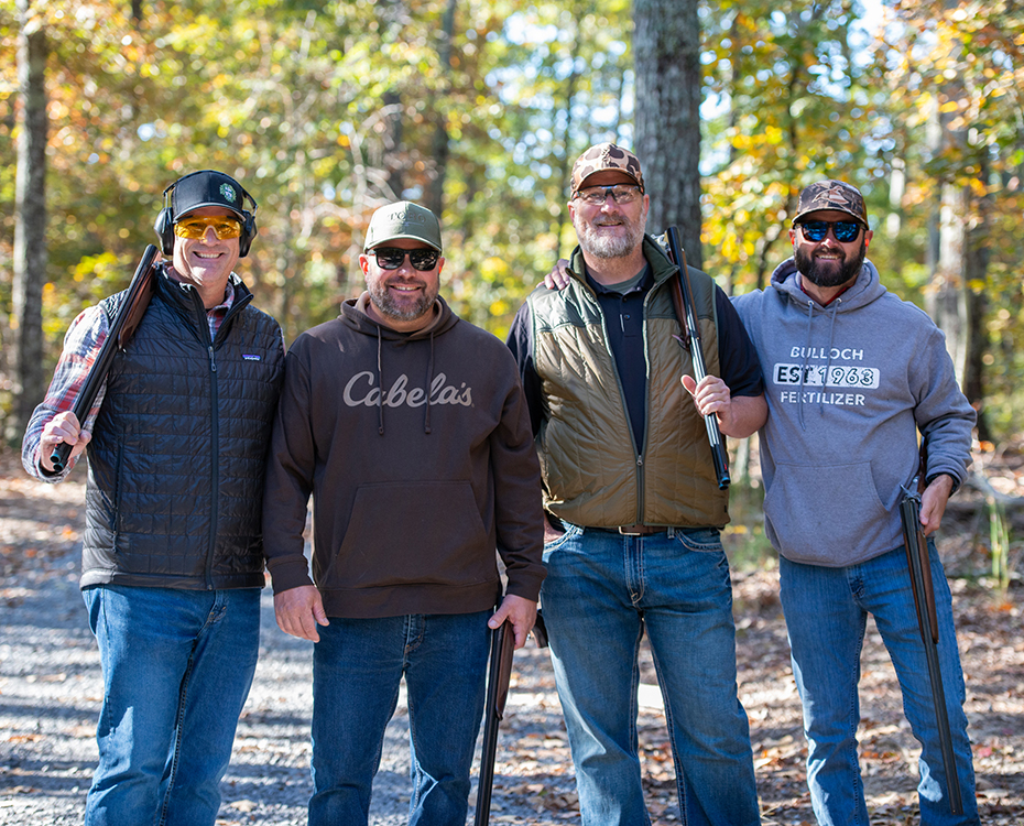 Four people standing outdoors in a forested area, wearing casual clothing including hats, jackets, and sunglasses. They are holding rifles and smiling, with trees and dappled sunlight in the background.