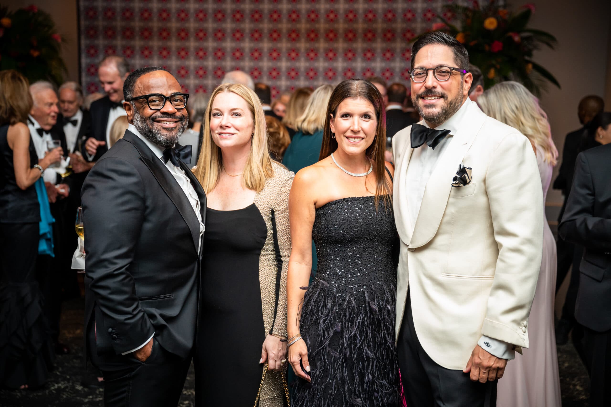Four people dressed formally pose together at an event. The background shows other attendees mingling and decorative elements with red and white patterns.