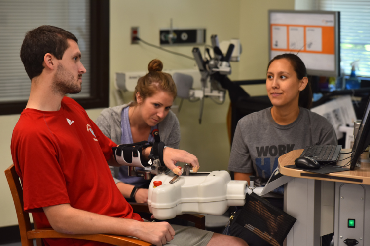 A man interacts with a device in a lab setting, assisted by two women. They are seated around a table with a computer screen displaying graphs and charts in the background.