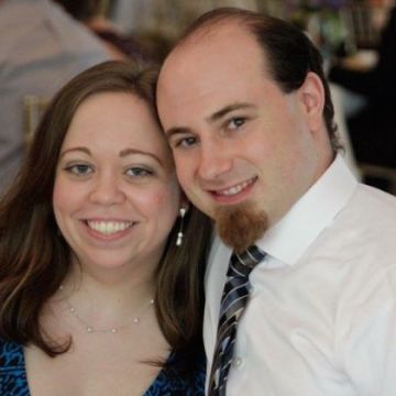 A man and woman smiling together. The woman has long brown hair and is wearing a blue top and earrings. The man has short hair and a goatee, wearing a white shirt and striped tie. They are at an indoor event with people in the background.