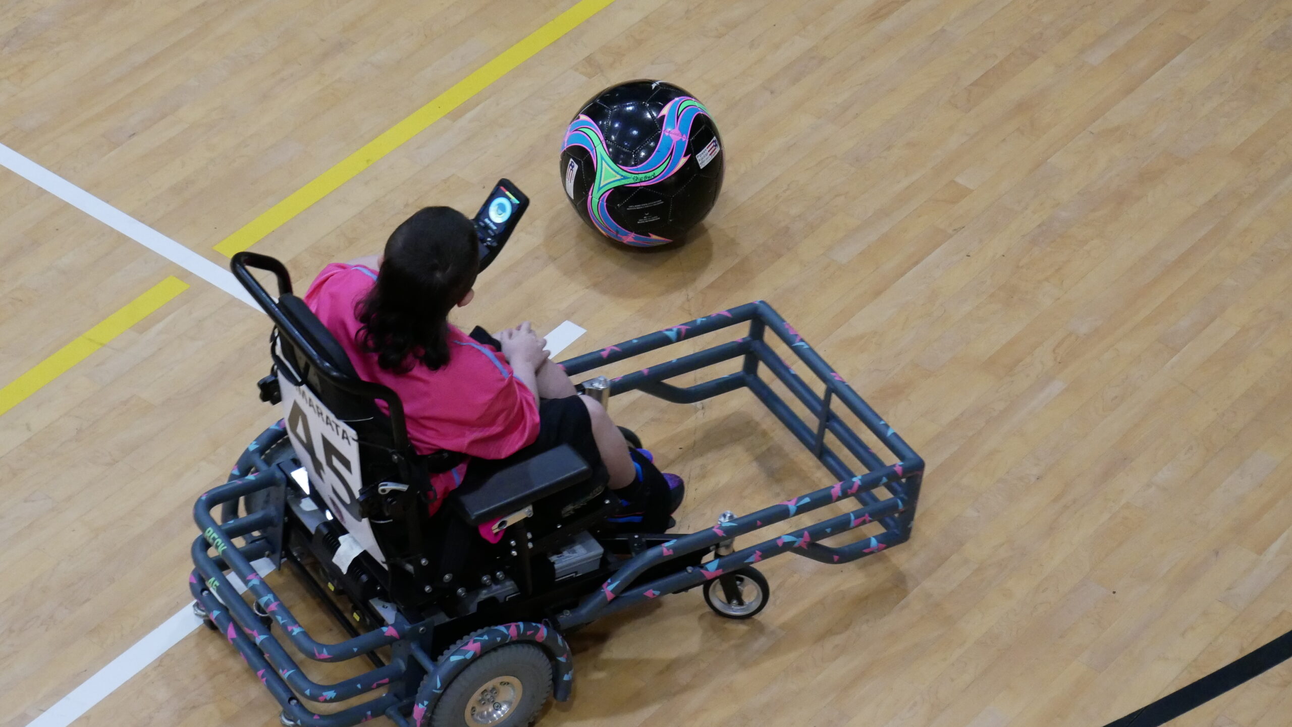 A person in a motorized wheelchair uses a mounted controller to play with a large, decorated black ball on a gymnasium floor.