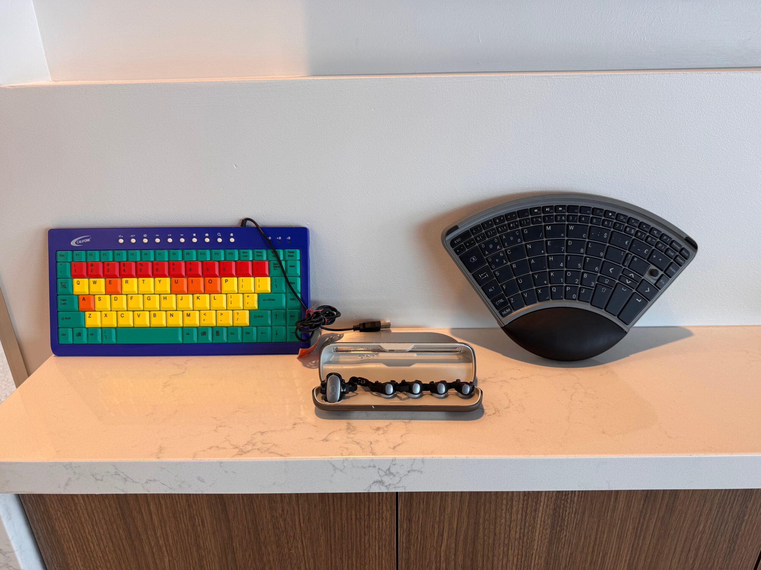 A colorful keyboard with large keys, a curved black ergonomic keyboard, and a small device with black knobs in a transparent case are displayed on a white countertop.