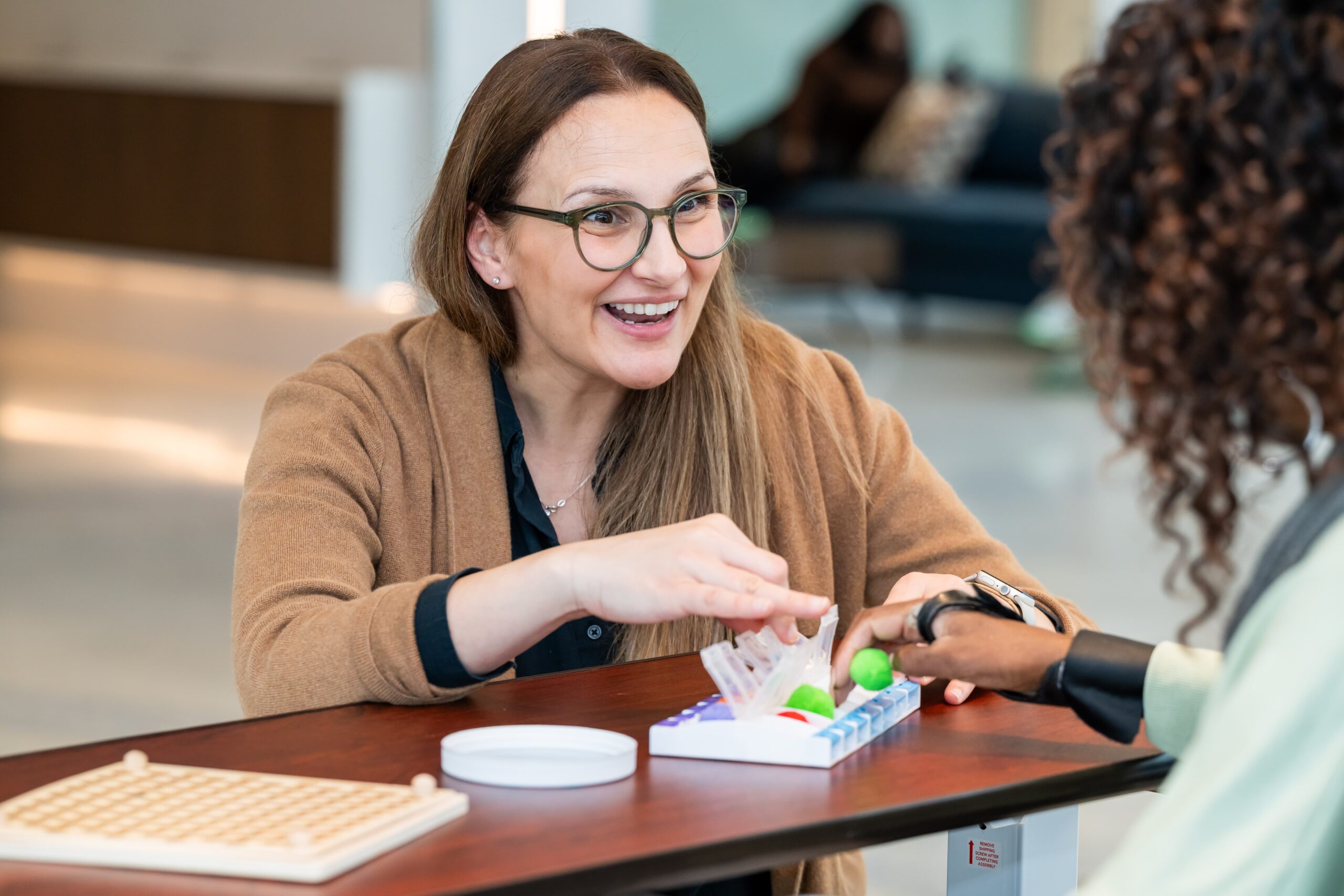 A woman with glasses, smiling, sits at a table and plays a board game with another person. She is reaching for a piece on the game board, and the setting appears to be a casual indoor space.