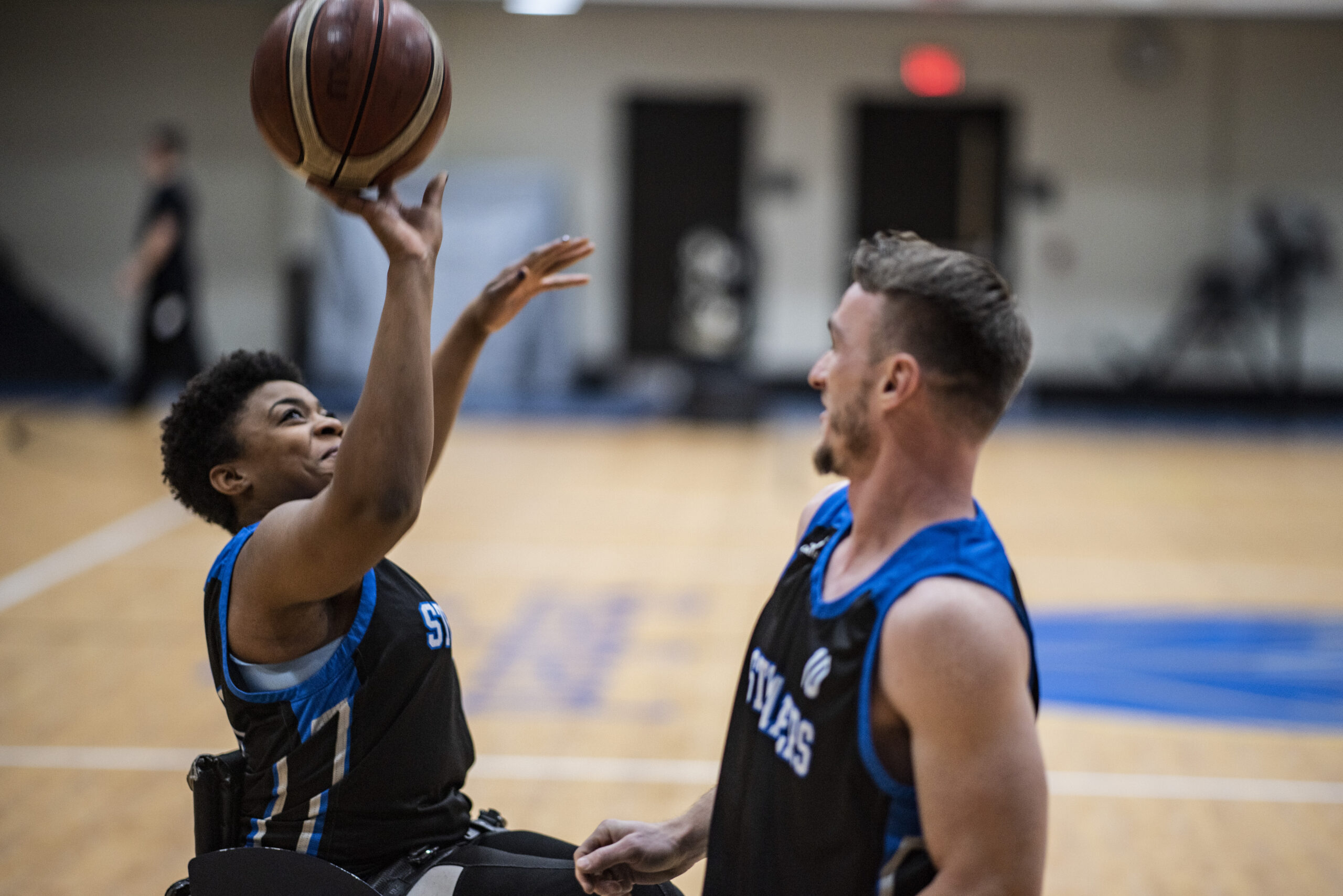 Two wheelchair basketball players in black and blue uniforms smile as one shoots the ball towards the hoop while the other looks on during a game in an indoor gym.