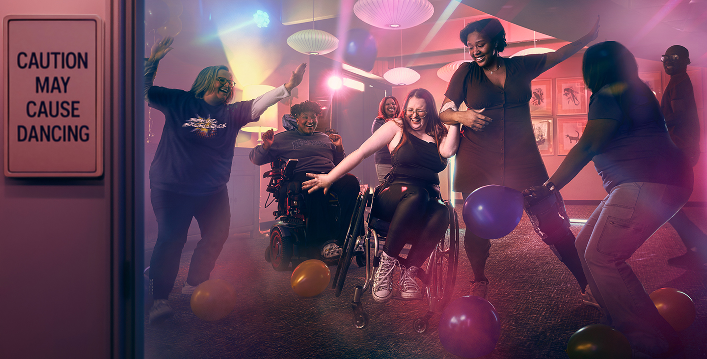 A diverse group of people, including wheelchair users, joyfully dance and play with balloons in a neon-lit room. A sign on the door reads, “Caution: May Cause Dancing.” The atmosphere is energetic and inclusive.