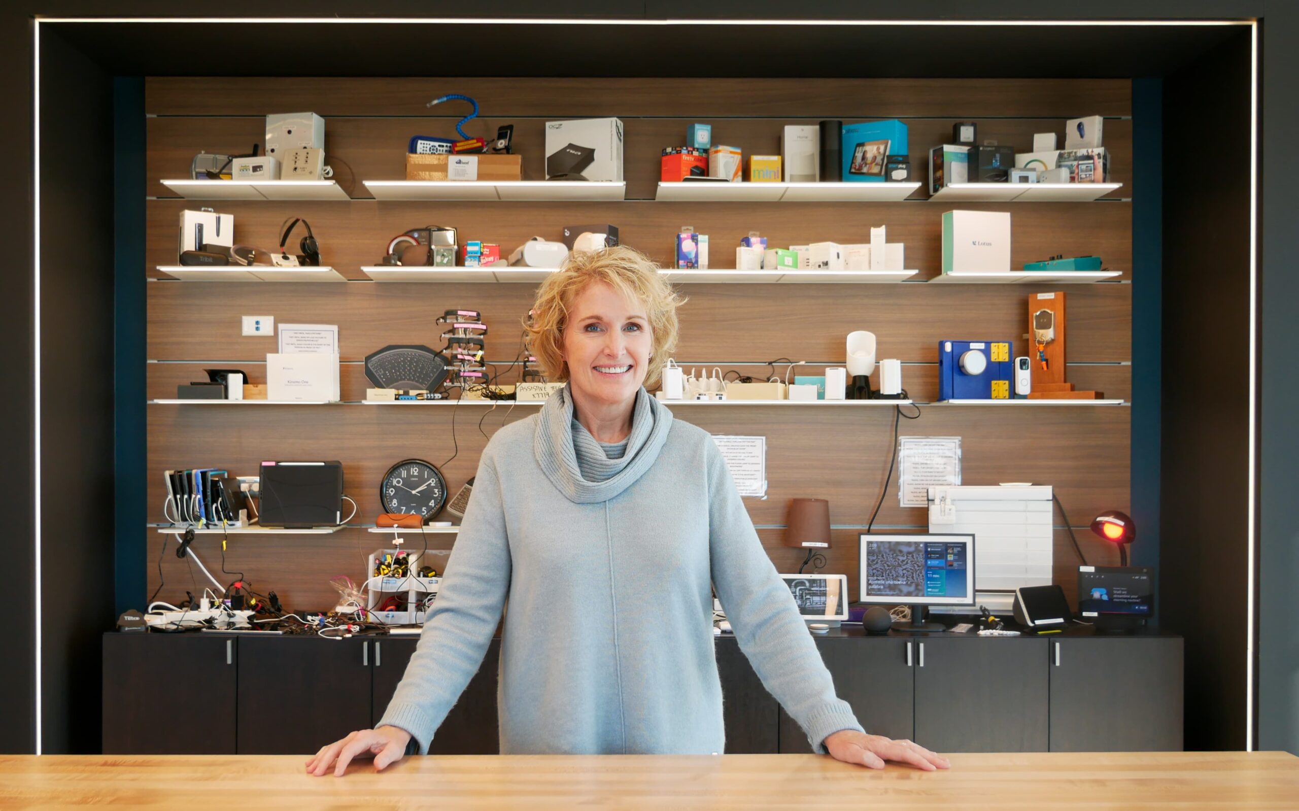 A woman stands smiling behind a wooden counter with shelves full of electronic gadgets, devices, and components on the wall behind her in a modern, tech-focused workspace.