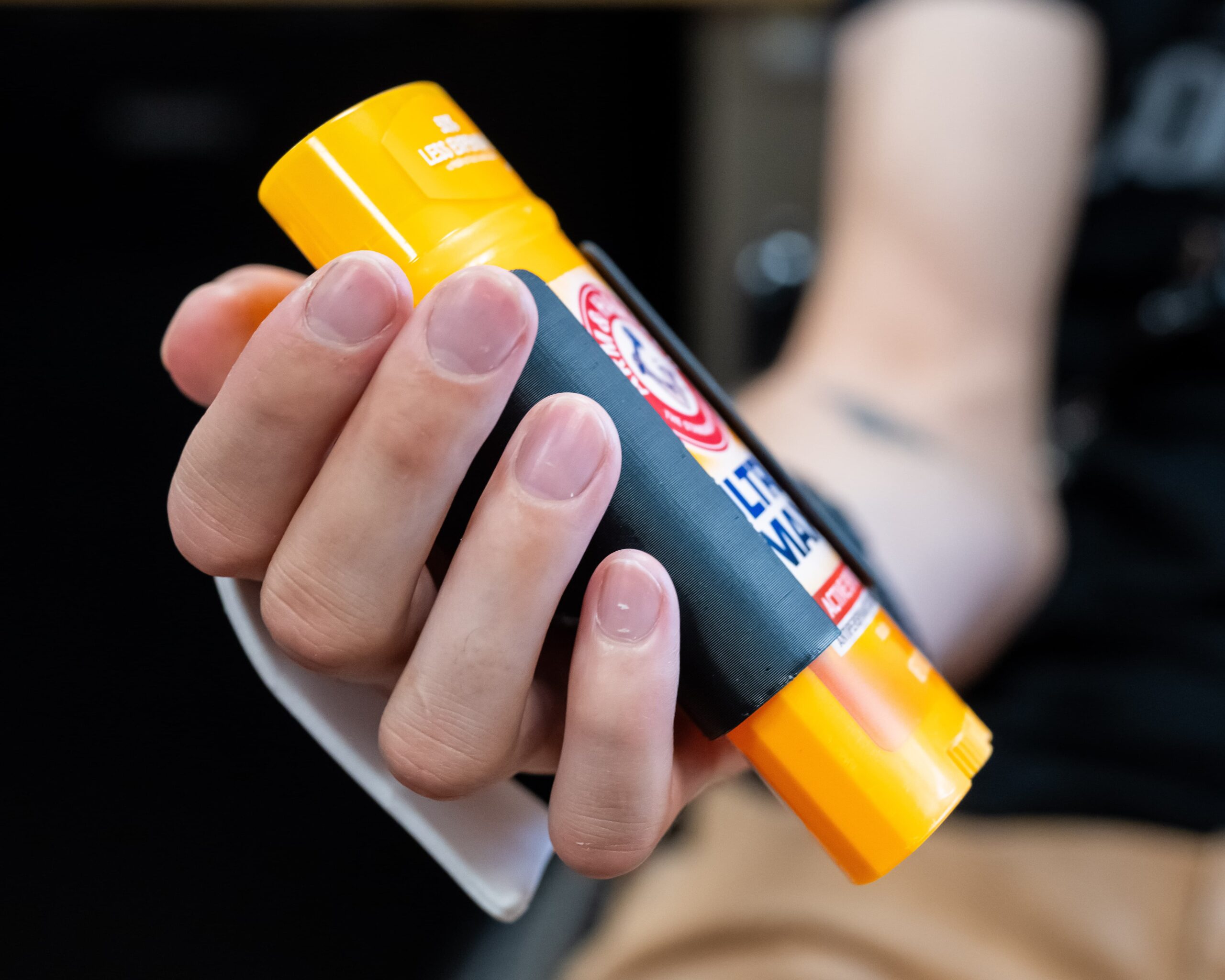 A close-up of a person’s hand holding a yellow plastic tube with a black label, possibly a stick of deodorant or lotion, with focus on the fingers and short nails.