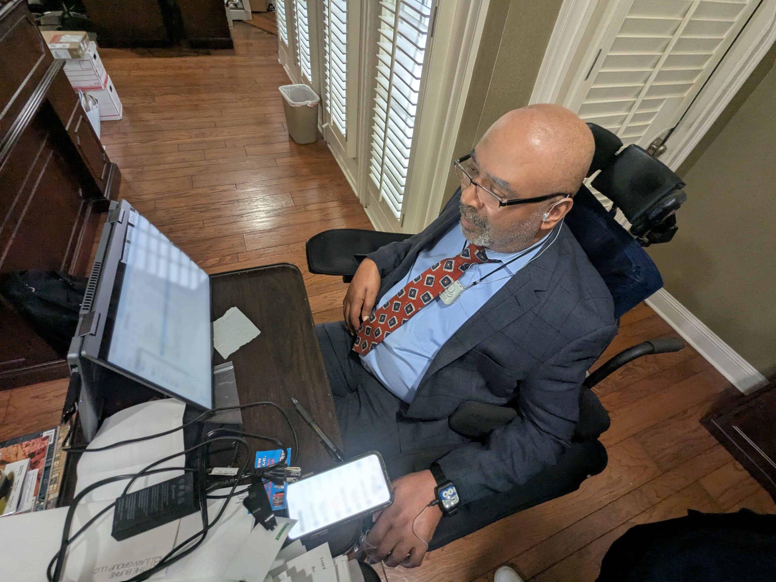 A man in a suit sits in a wheelchair at a desk, working on multiple monitors and a smartphone. He appears focused, with various office supplies and paperwork around him in a well-lit room with wooden floors.