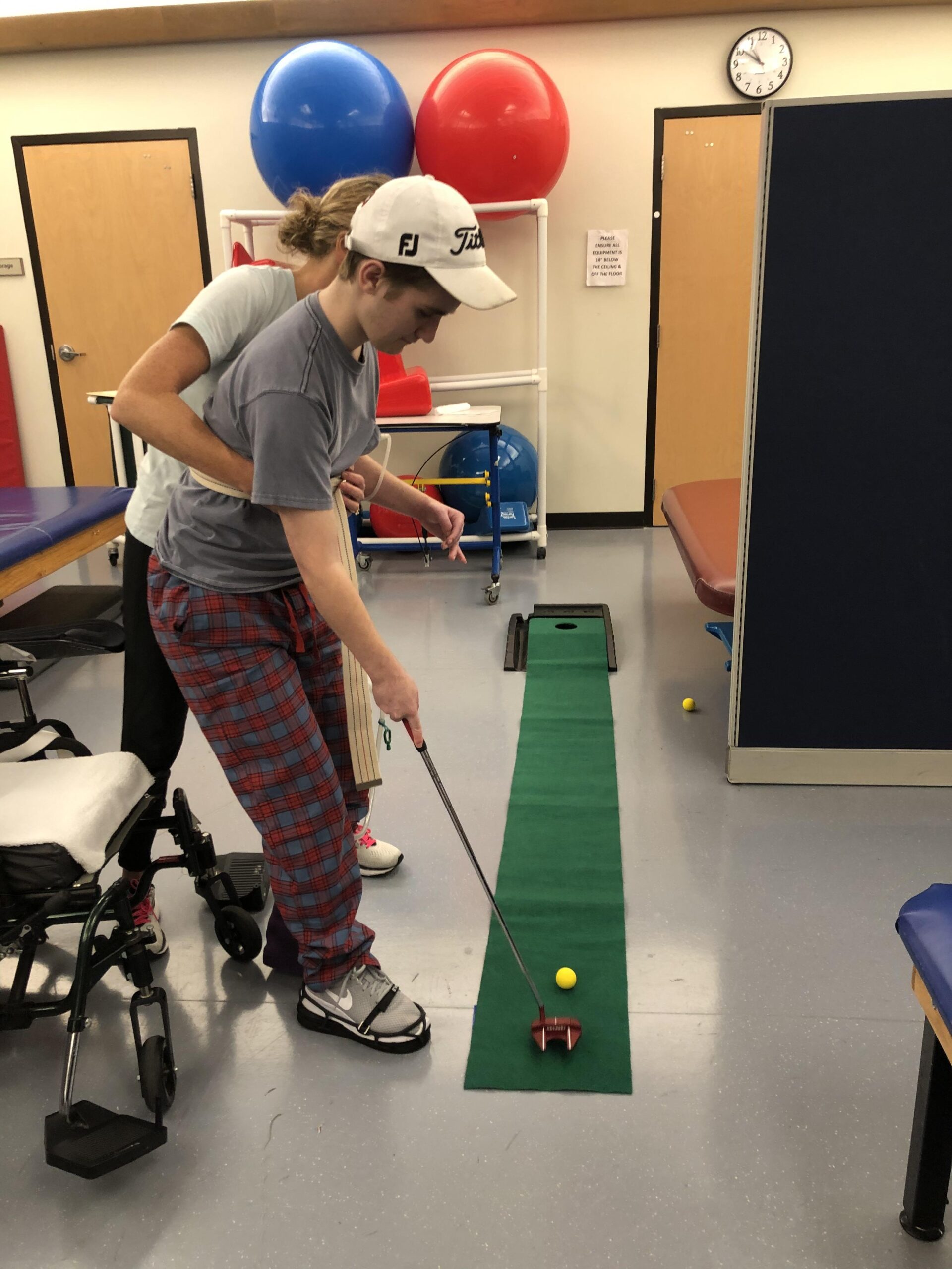 A therapist helps a patient practice putting on a small indoor green mat. Exercise balls sit in the background with a wheelchair nearby.