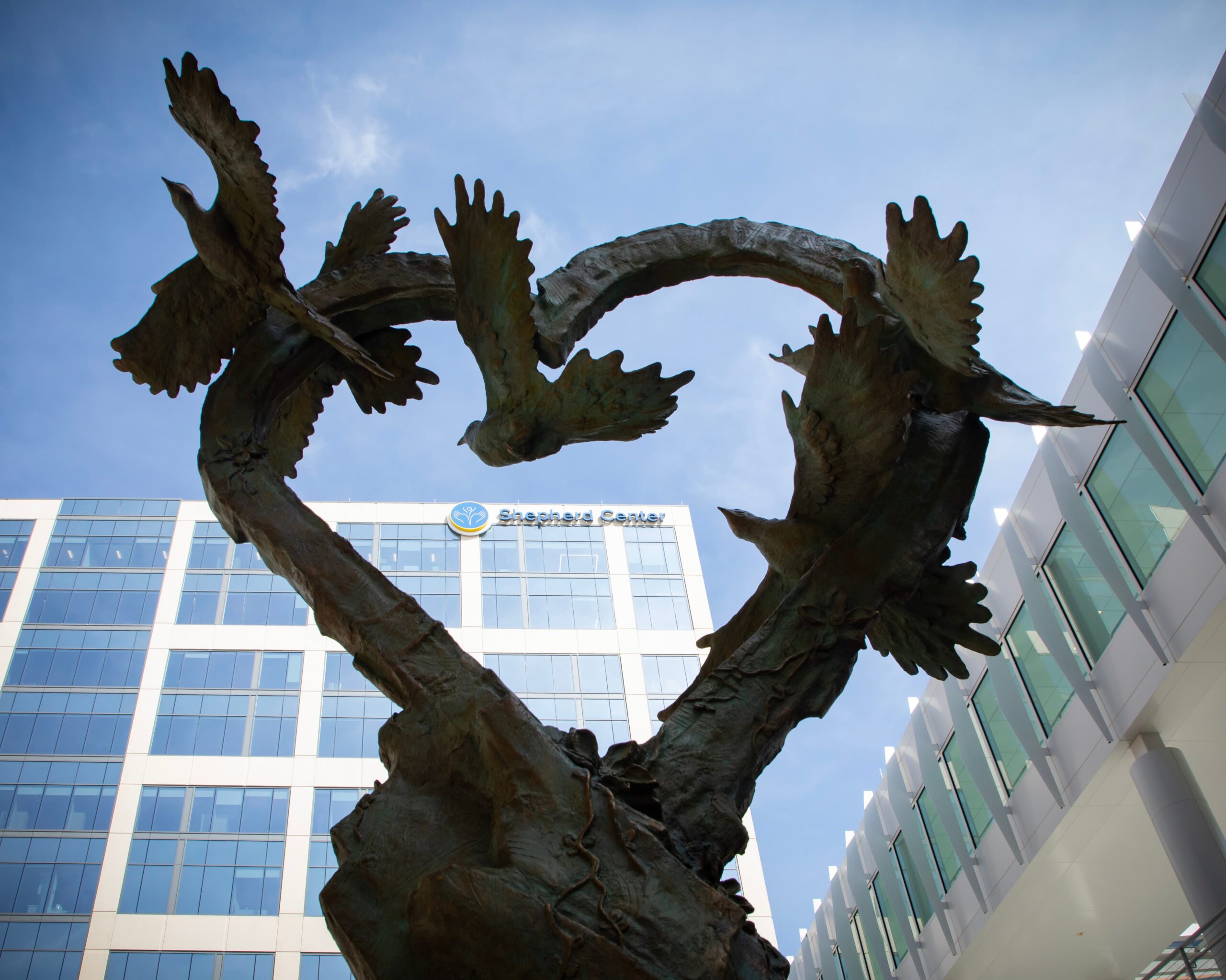 A bronze sculpture of two birds forming a heart shape, set against the backdrop of modern glass office buildings with a sign that reads 