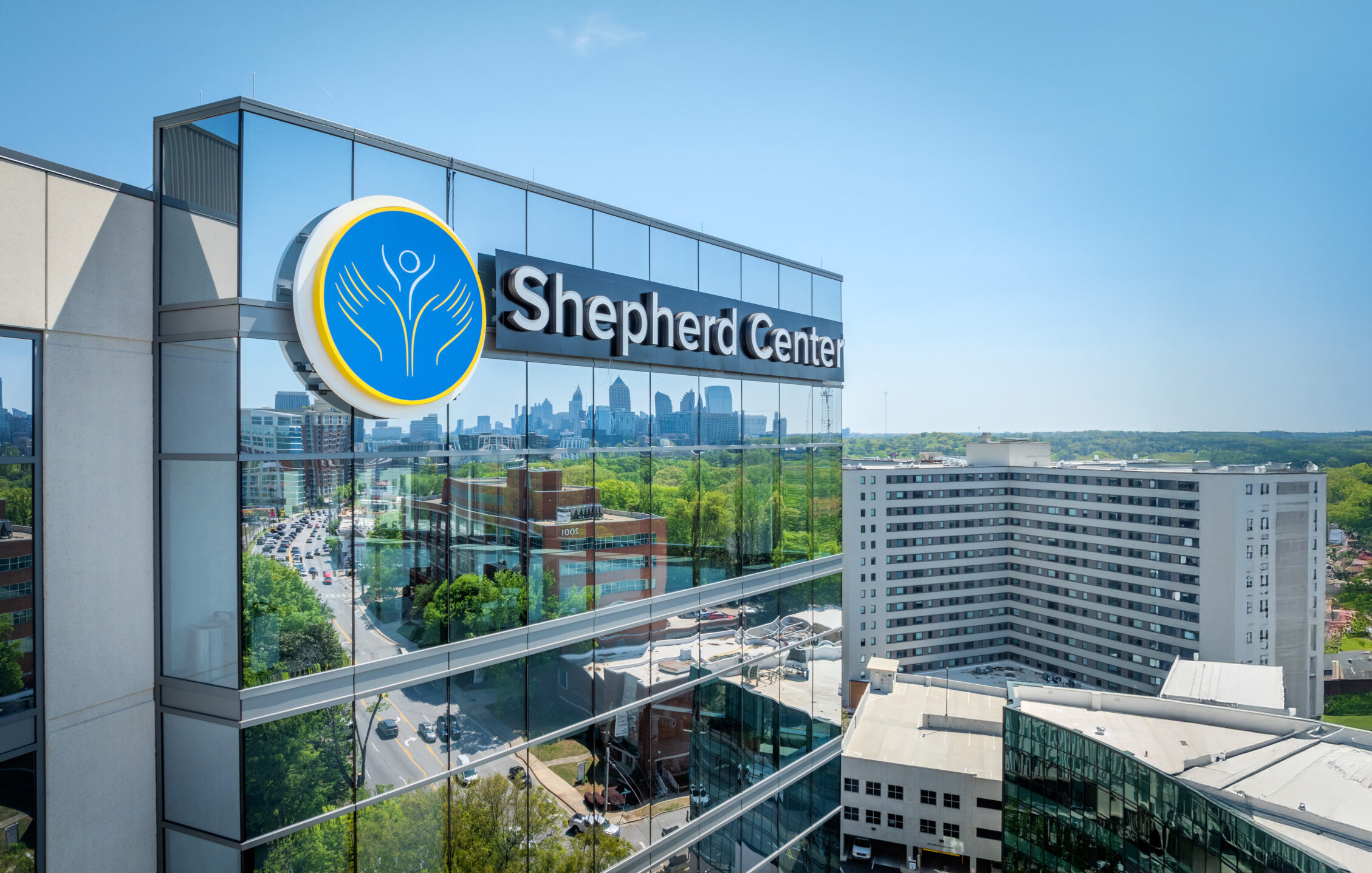 The exterior of the Shepherd Center building with its logo and name displayed prominently, reflecting the city skyline and greenery on its glass windows under a clear blue sky.