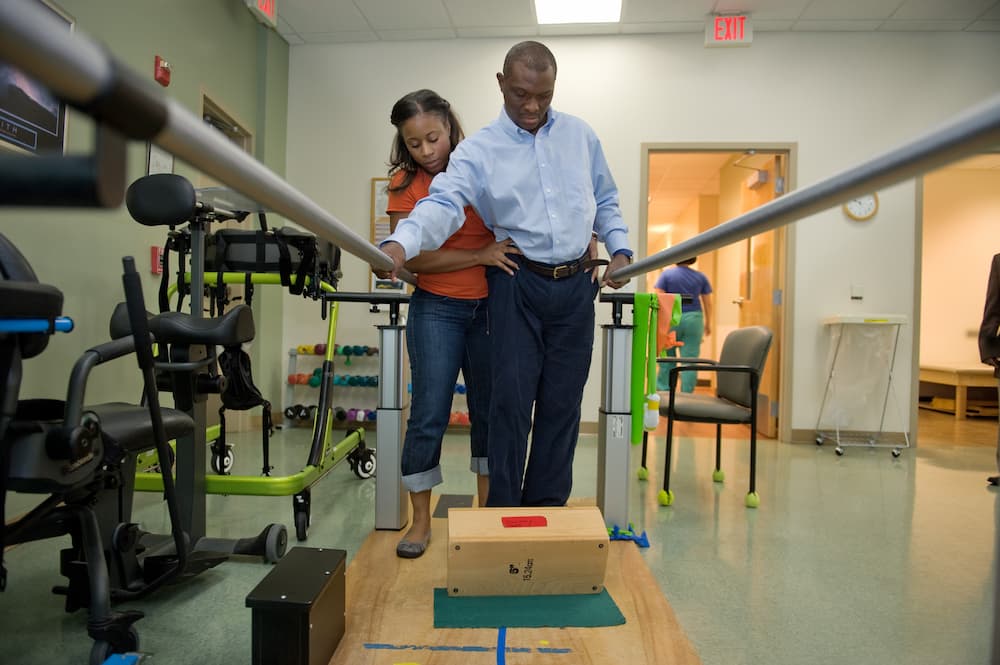 A physical therapist at Shepherd Center assists a man with walking between parallel bars in the rehabilitation room, which is equipped with mobility aids and exercise equipment.