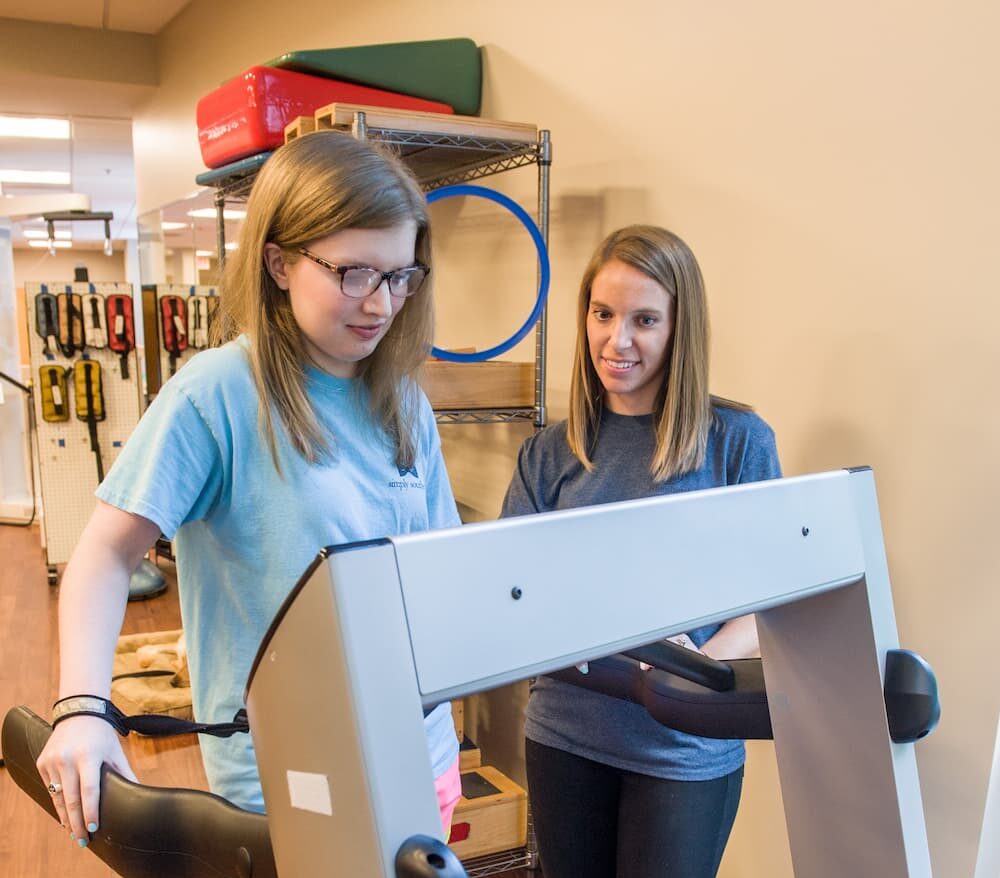 A woman walks on a treadmill under the watchful eye of another. Both are casually dressed, surrounded by exercise equipment and shelves.