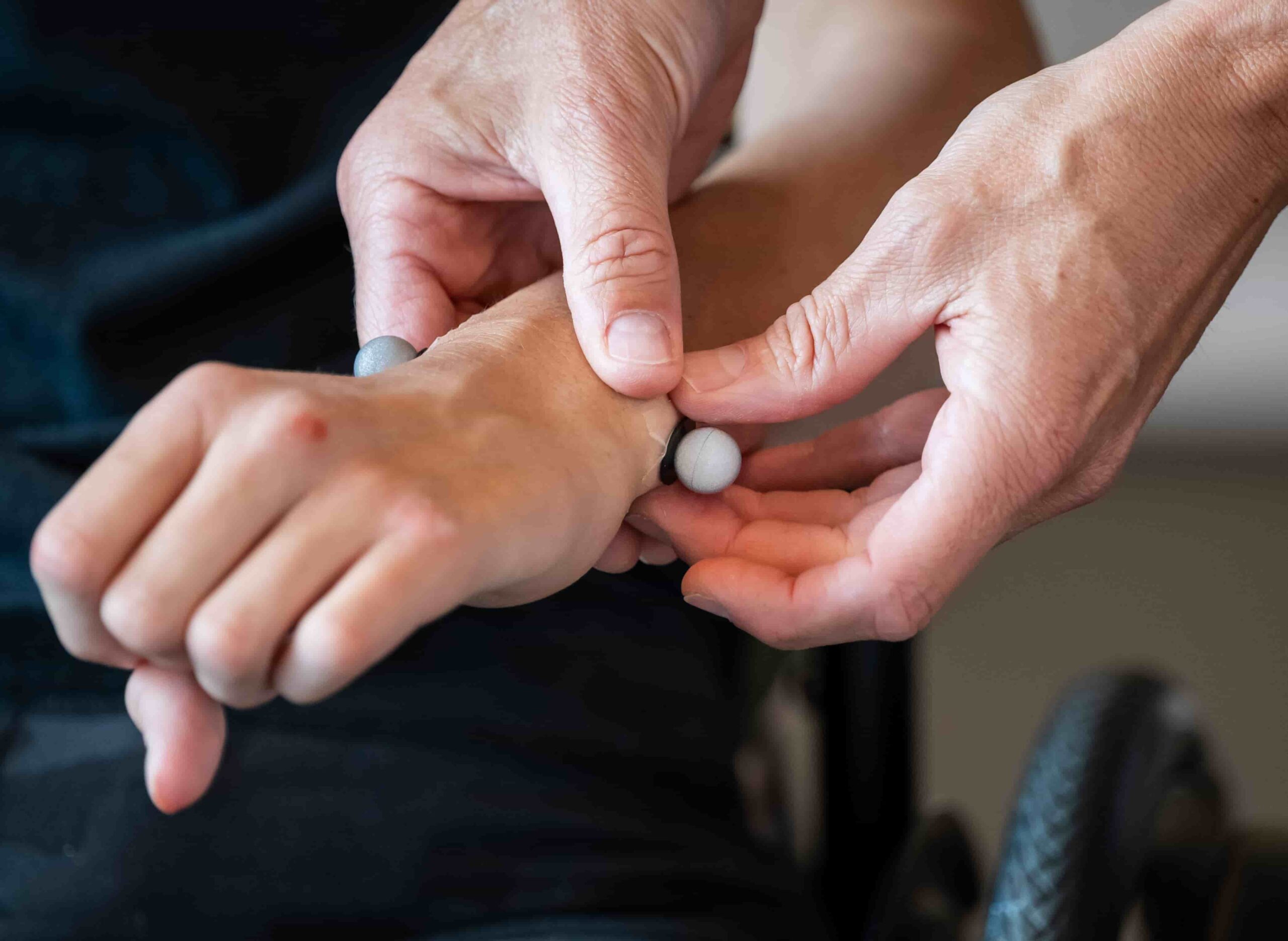 Close-up of a person’s hands pressing on another person's wrist in a therapeutic or medical setting. A wheelchair wheel is visible in the background.