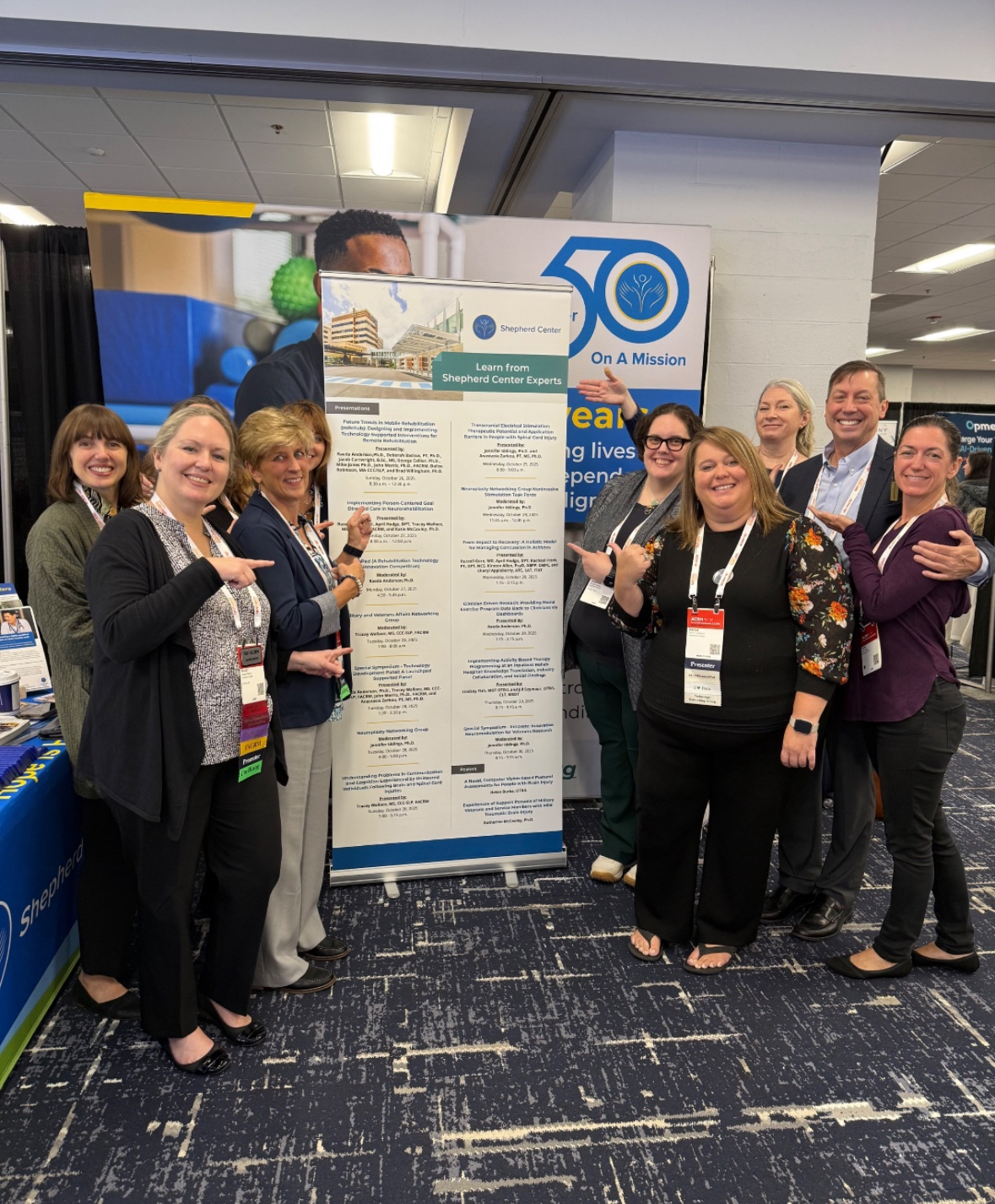 Nine smiling people stand indoors around a tall informational display banner with 