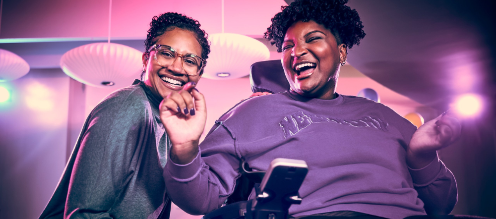 Two women, one wearing glasses and the other in a wheelchair, share laughter in a colorful, well-lit room. Their joyful connection highlights a sense of belonging as they enjoy being seen and valued in each other's company.