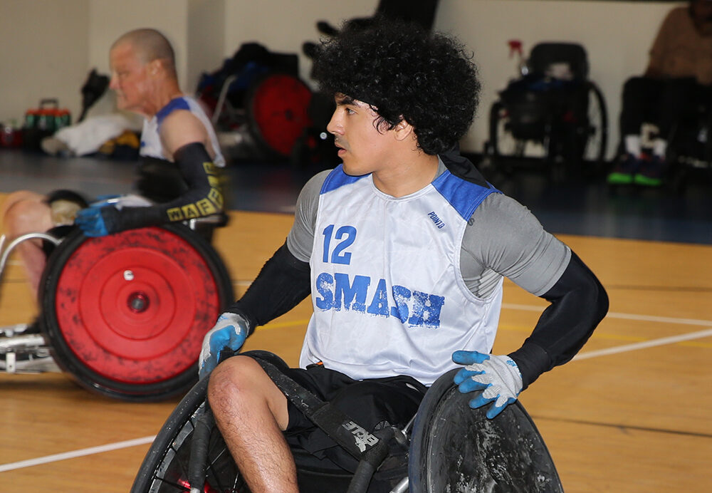 A young male athlete in a wheelchair, wearing a blue and white jersey with the number 12 and "SMASH" on the front, plays wheelchair rugby on an indoor court. Another player is visible in the background.