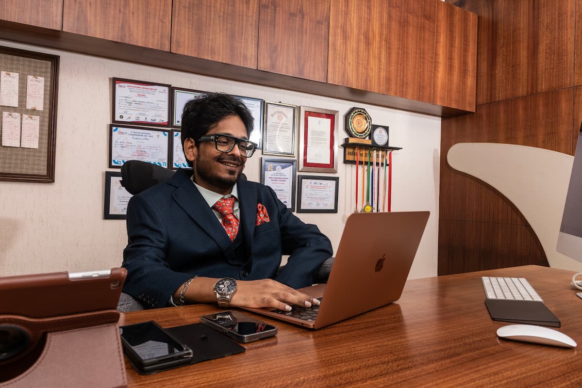 A man in a suit sits at a wooden desk, smiling at his laptop. Behind him, framed certificates and awards are displayed on the wall. The desk has a keyboard, tablet, phone, and other devices.