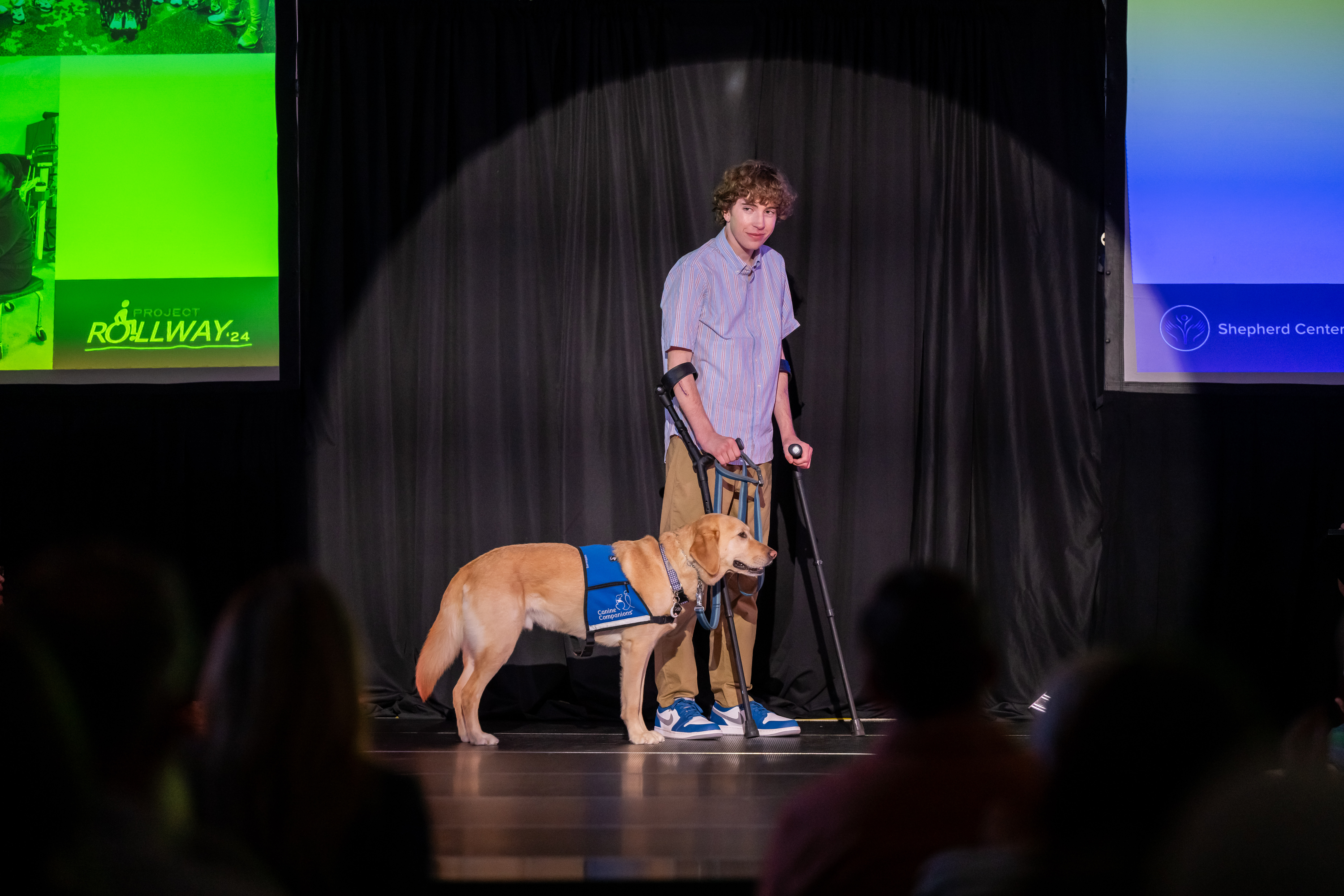 A person on crutches stands on a stage next to a service dog wearing a blue vest. They are under a spotlight, with a dimly lit audience and screens in the background displaying logos.
